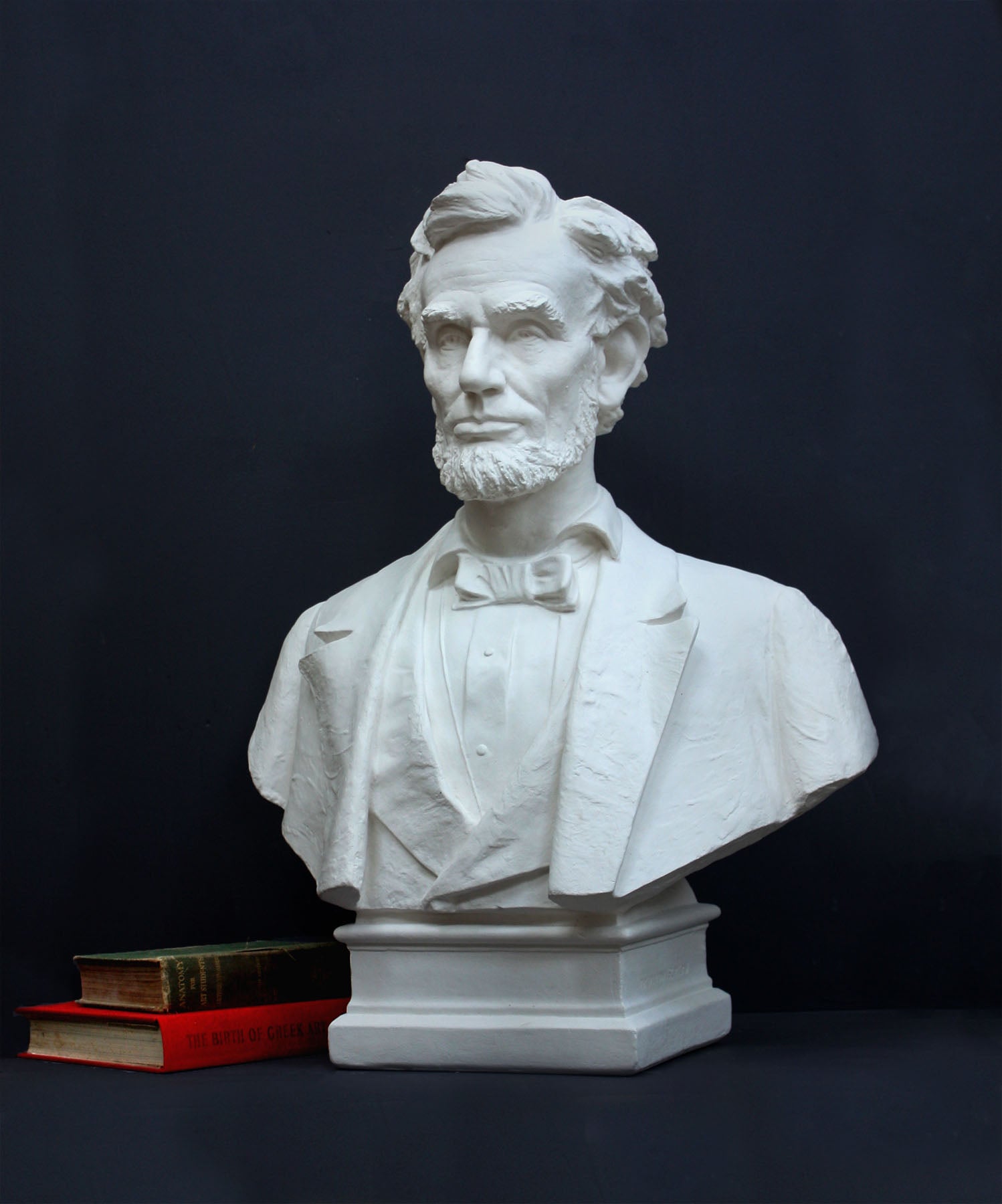 image of a plaster sculpture bust of man, namely Abraham Lincoln, next to stacked green and red books on a gray background