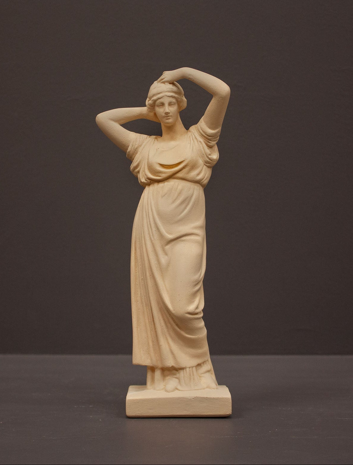 photo of off-white plaster cast statue of a female in robes with her arms raised to her head against gray background