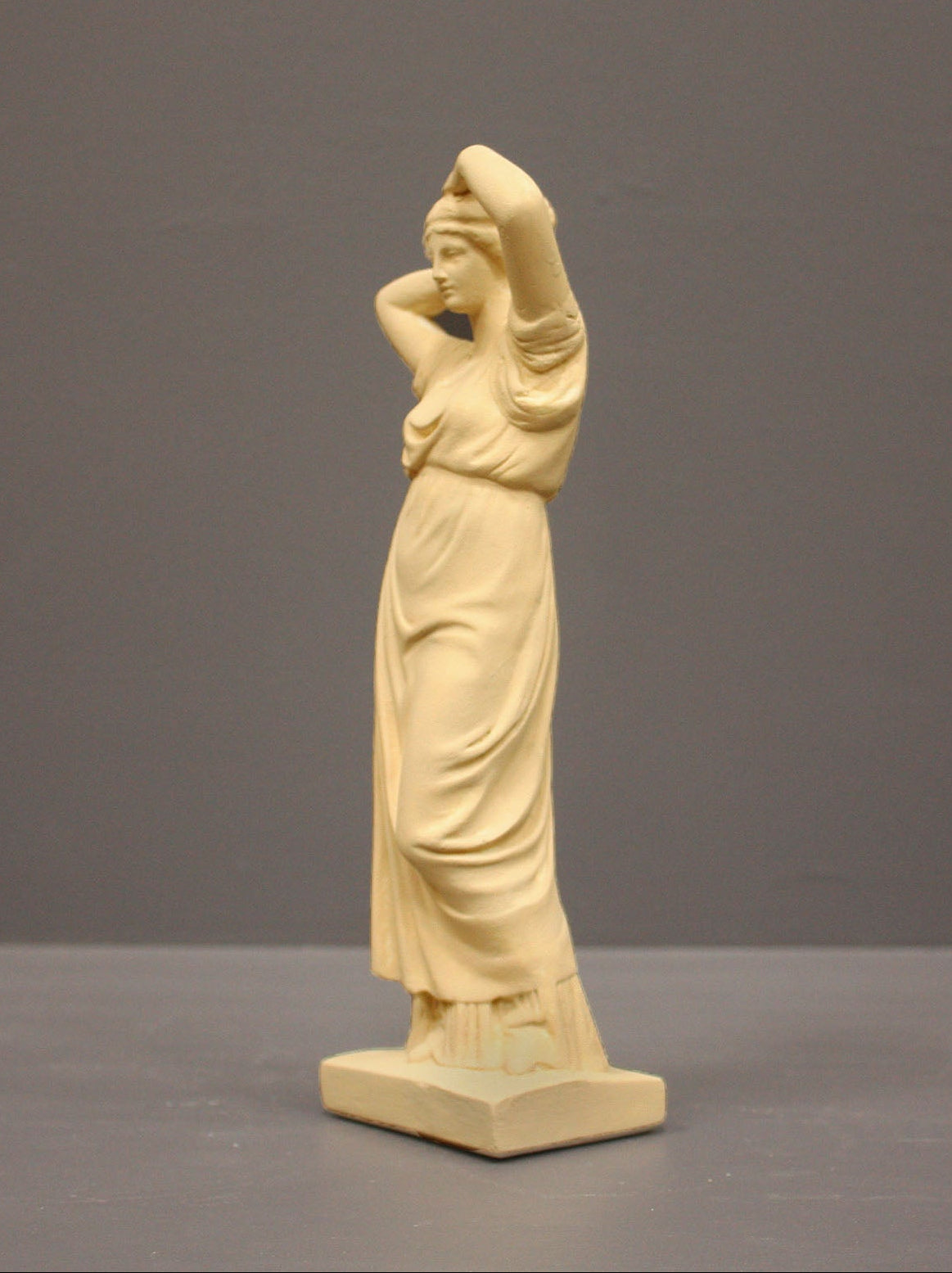 photo of off-white plaster cast statue of a female in robes with her arms raised to her head against gray background