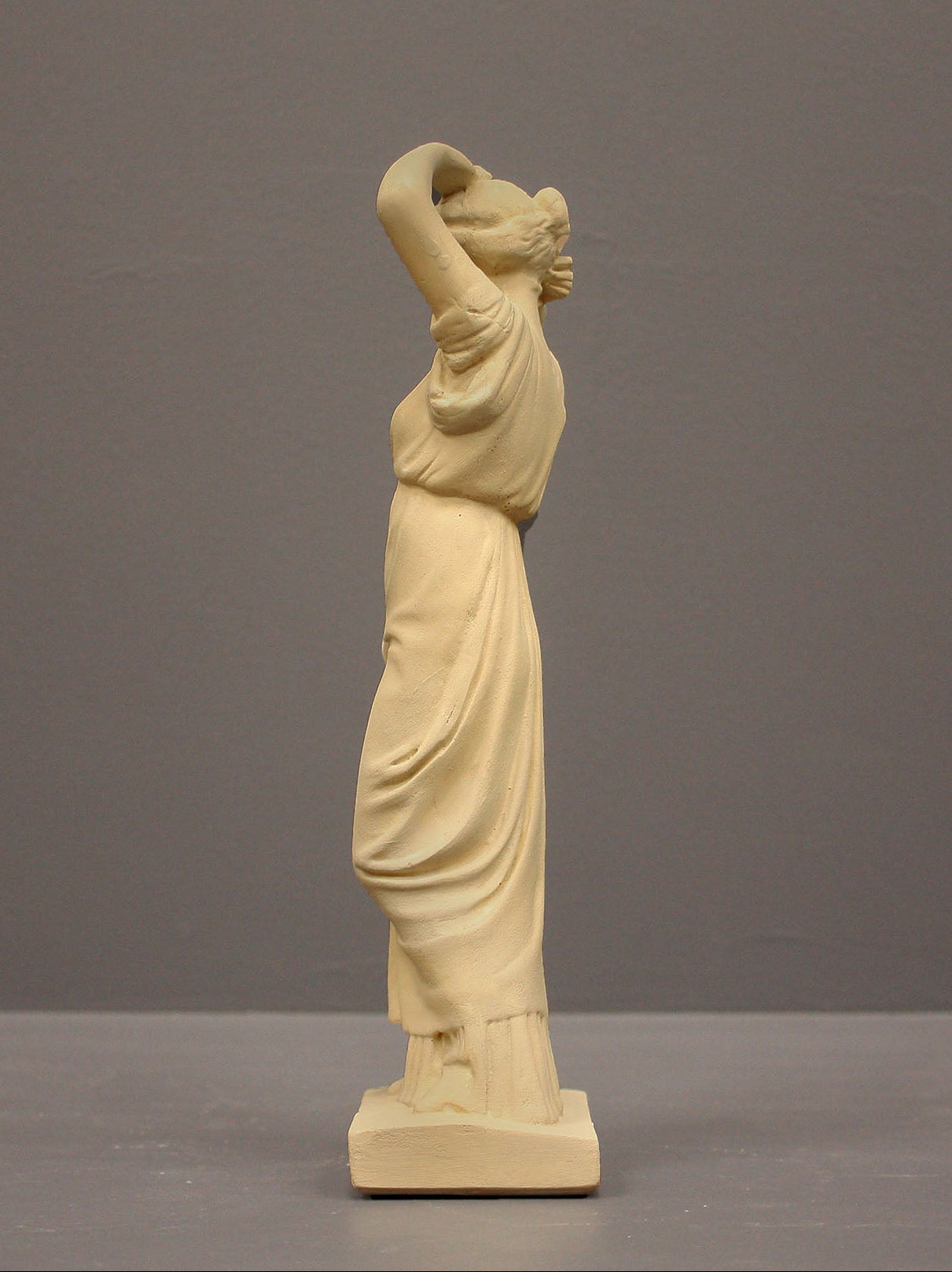 photo of off-white plaster cast statue of a female in robes with her arms raised to her head against gray background