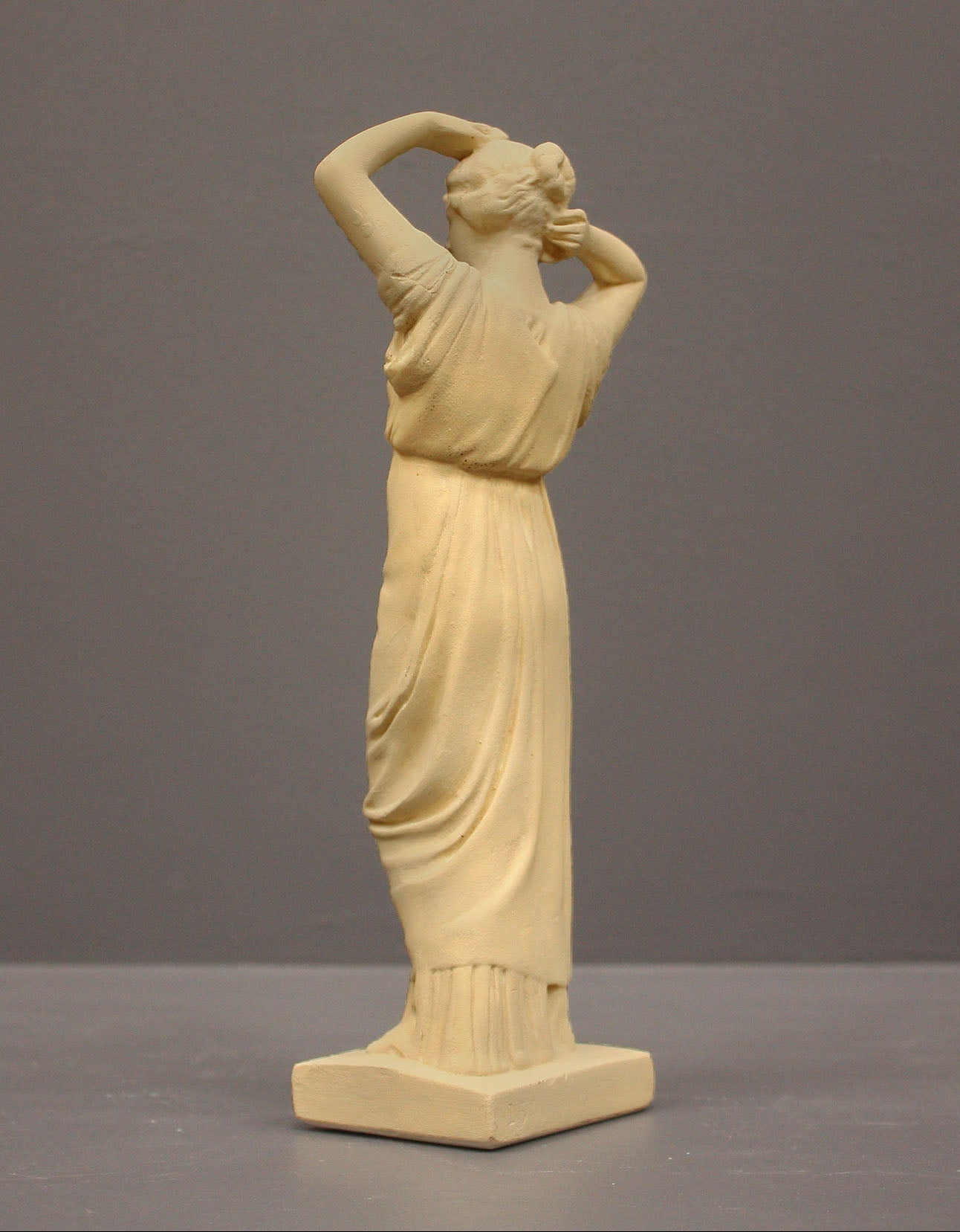 photo of off-white plaster cast statue of a female in robes with her arms raised to her head against gray background
