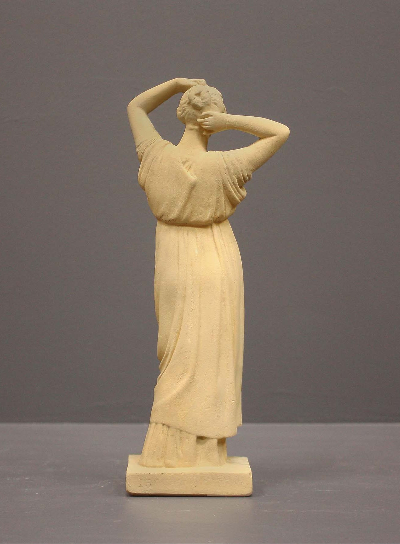 photo of off-white plaster cast statue of a female in robes with her arms raised to her head against gray background