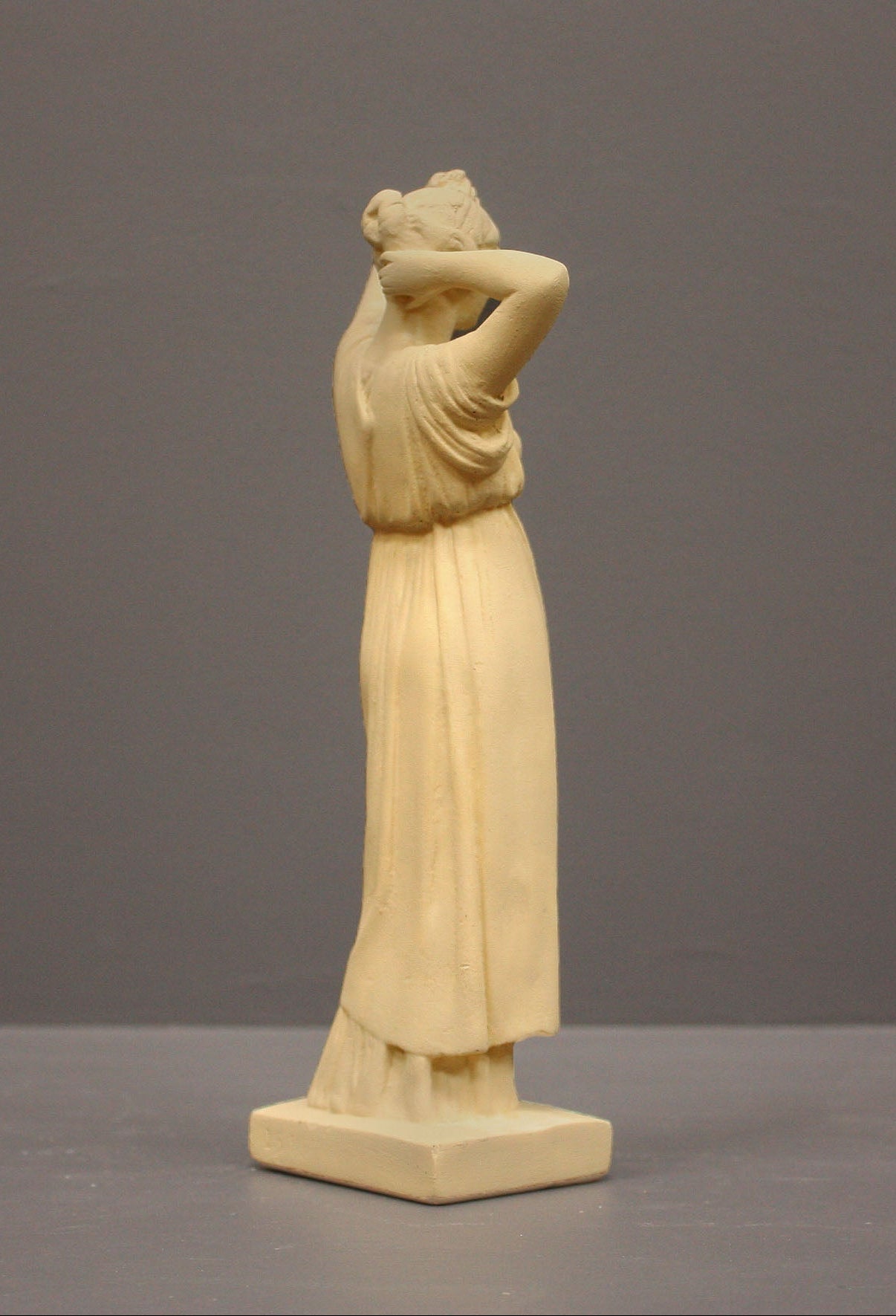 photo of off-white plaster cast statue of a female in robes with her arms raised to her head against gray background