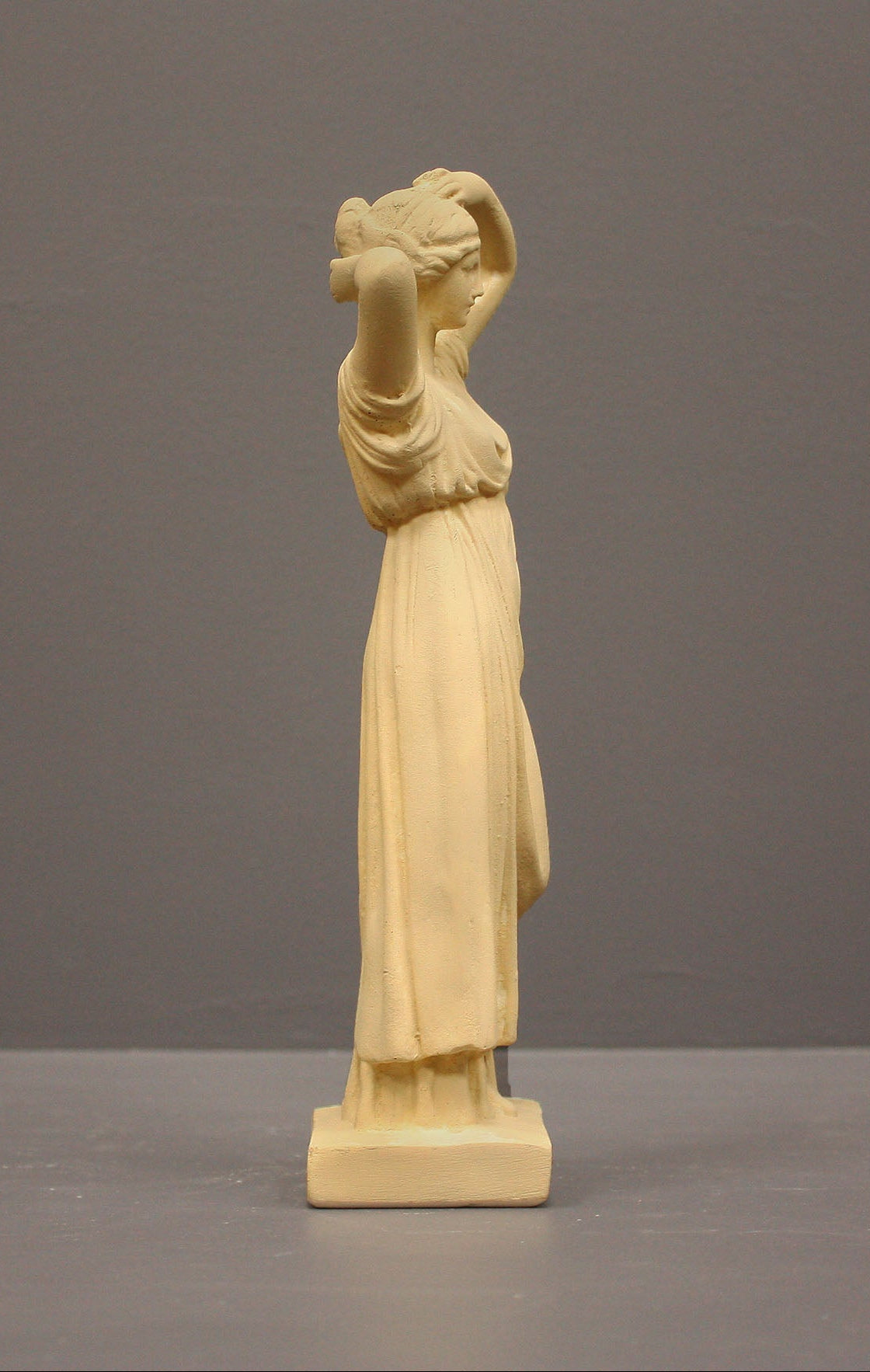 photo of off-white plaster cast statue of a female in robes with her arms raised to her head against gray background