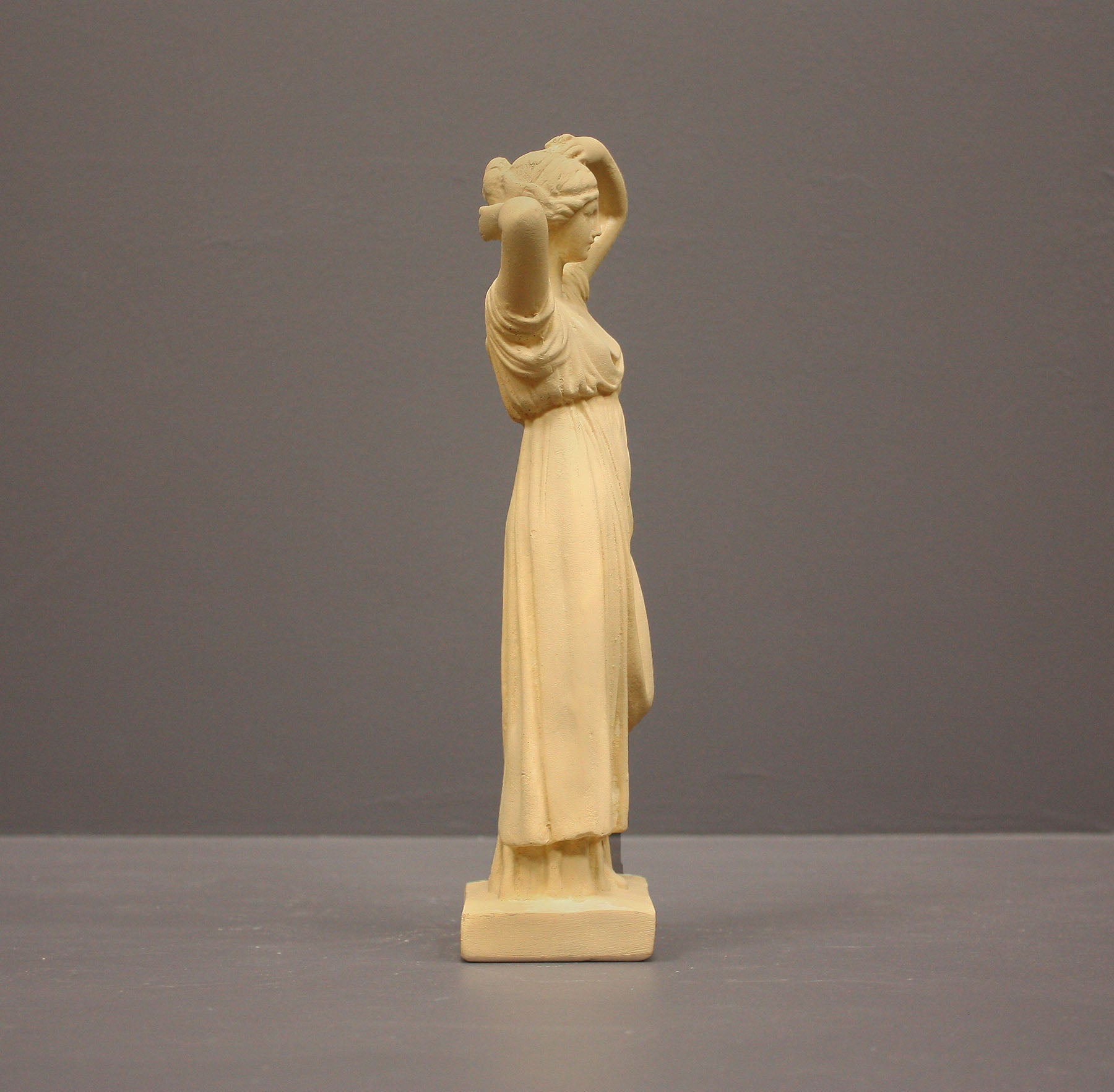 photo of off-white plaster cast statue of a female in robes with her arms raised to her head against gray background