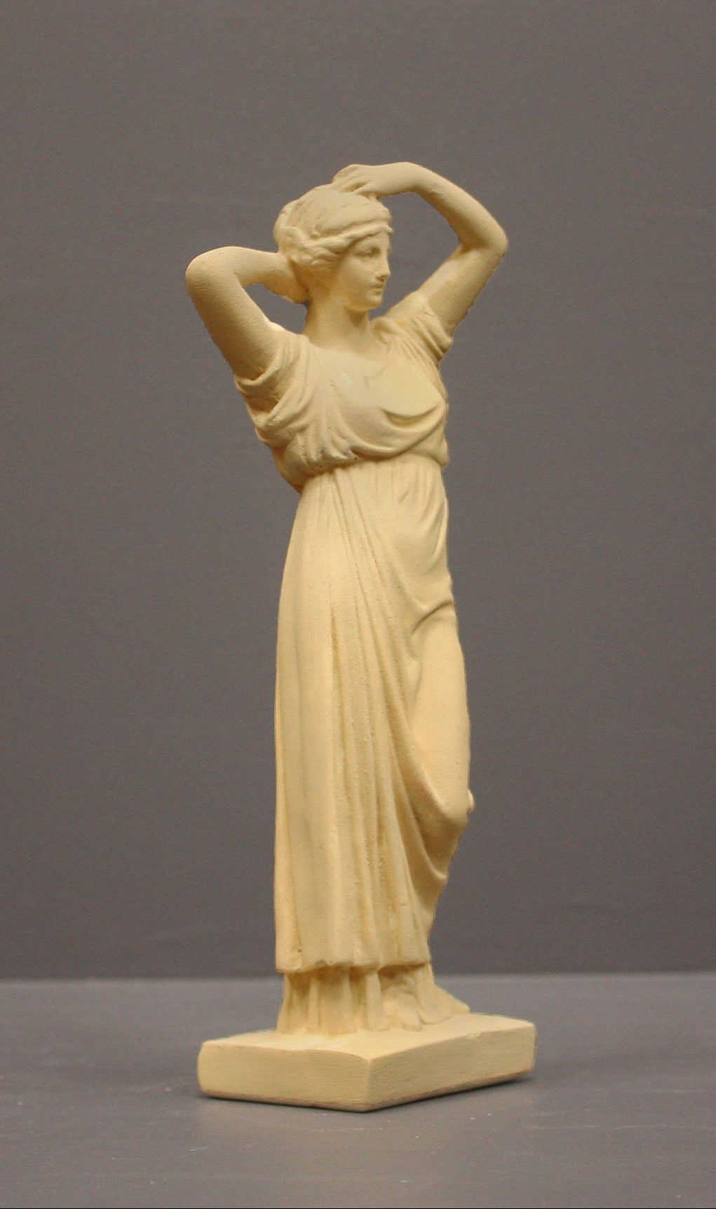 photo of off-white plaster cast statue of a female in robes with her arms raised to her head against gray background