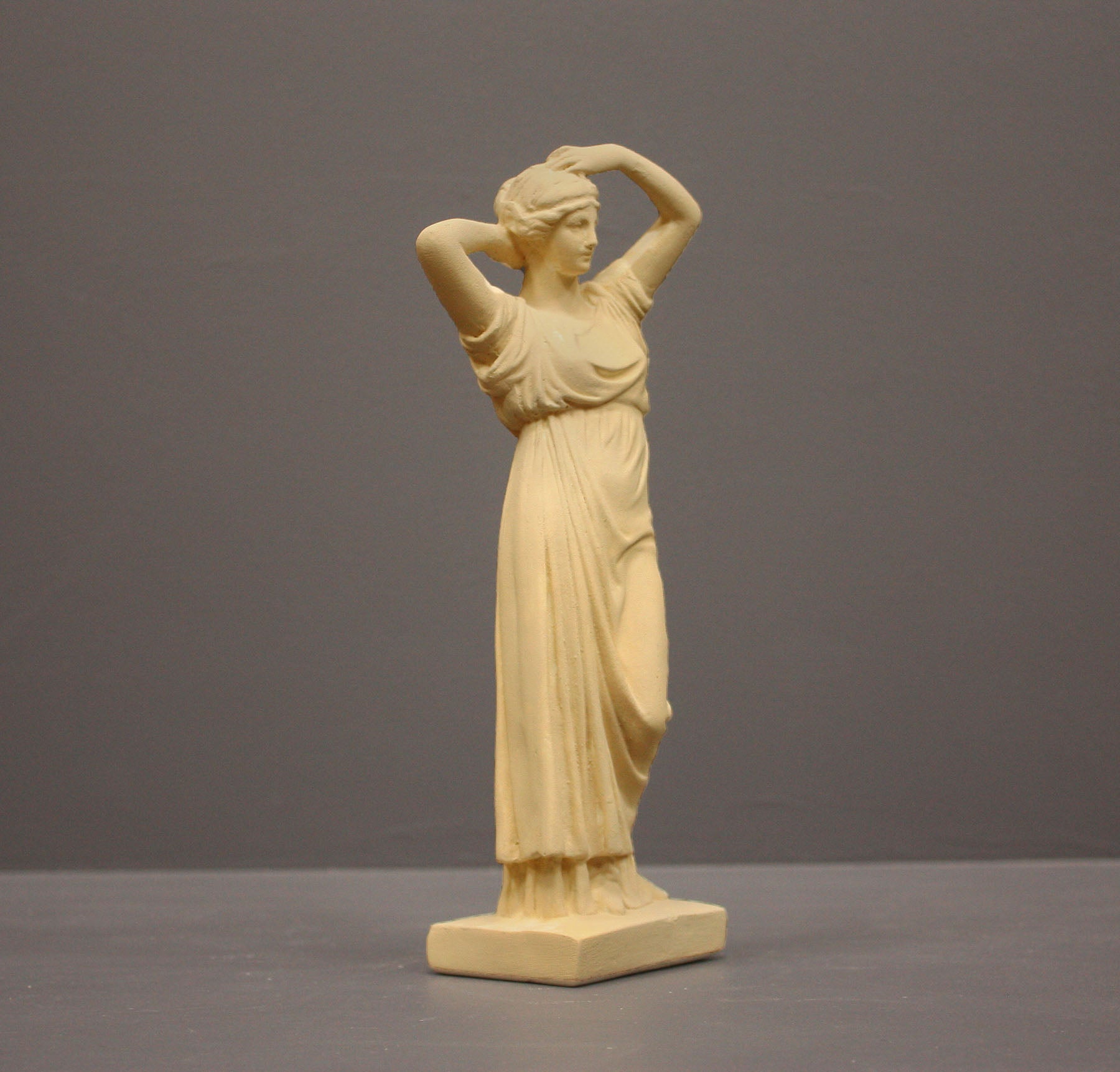 photo of off-white plaster cast statue of a female in robes with her arms raised to her head against gray background