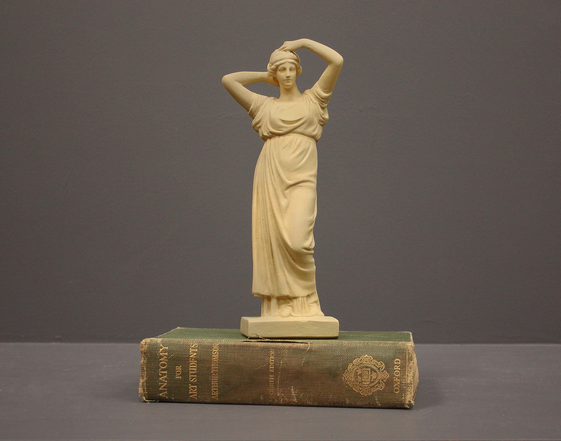 photo of off-white plaster cast statue of a female in robes with her arms raised to her head atop green book against gray background