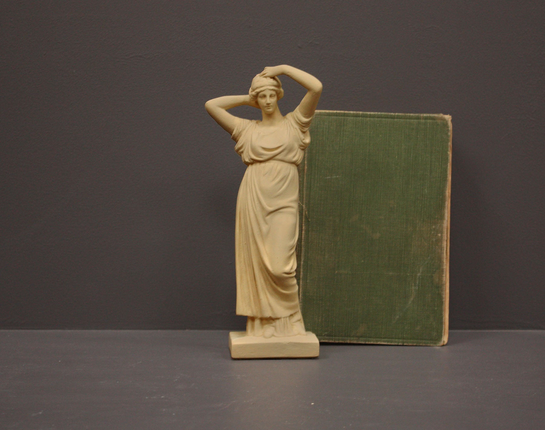 photo of off-white plaster cast statue of a female in robes with her arms raised to her head beside green book against gray background