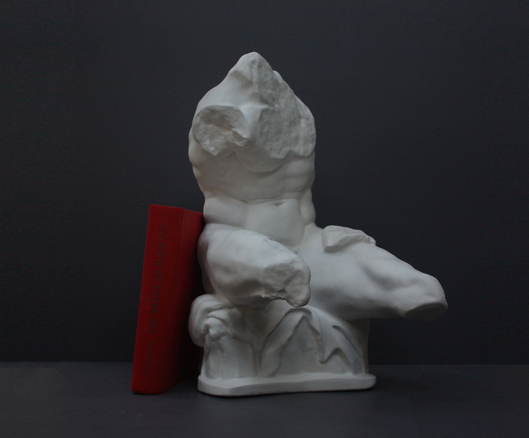 image of a plaster sculpture of nude male torso next to a red book on a gray background