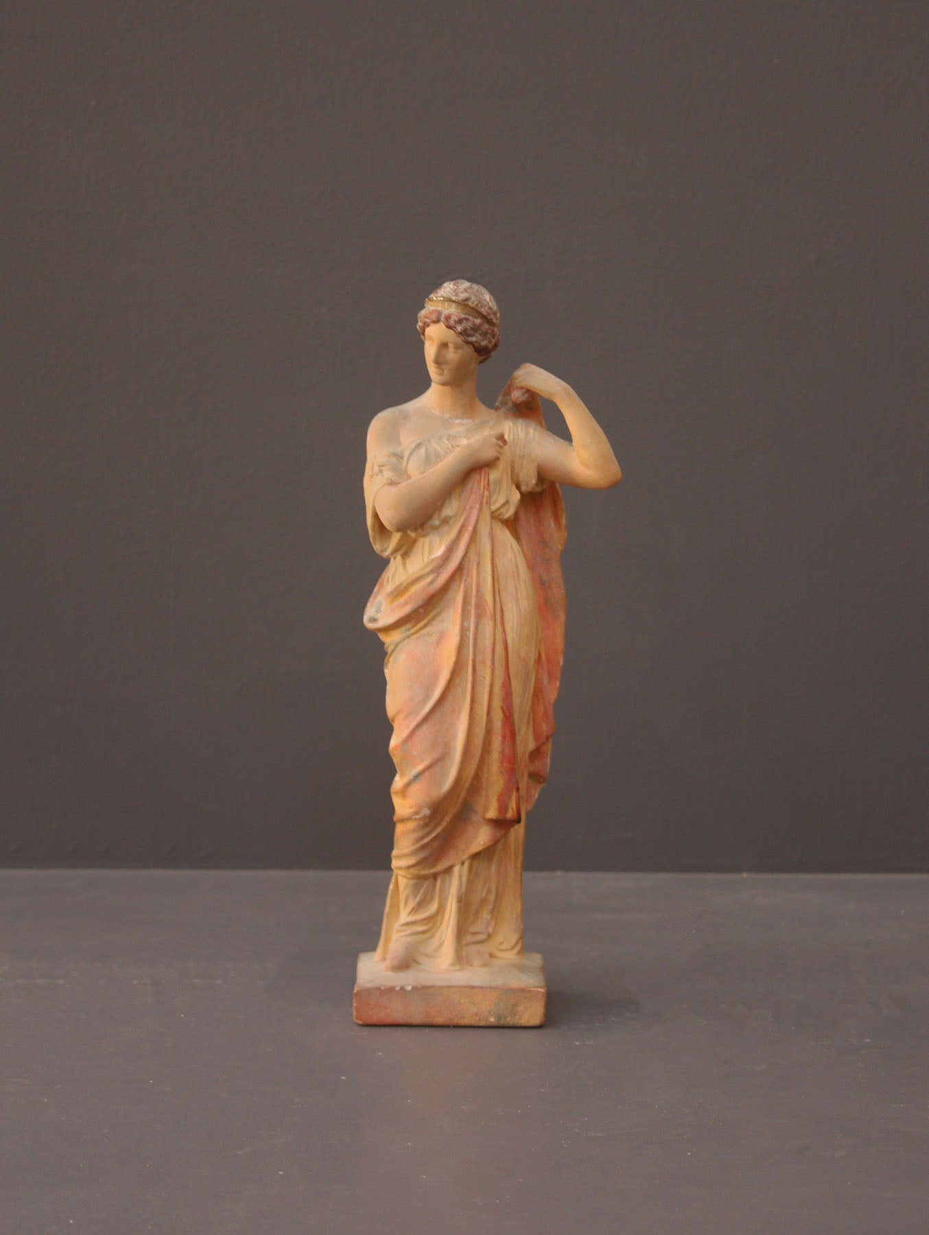 photo of plaster cast statue of female in pink robes with left arm raised and holding robe on gray background