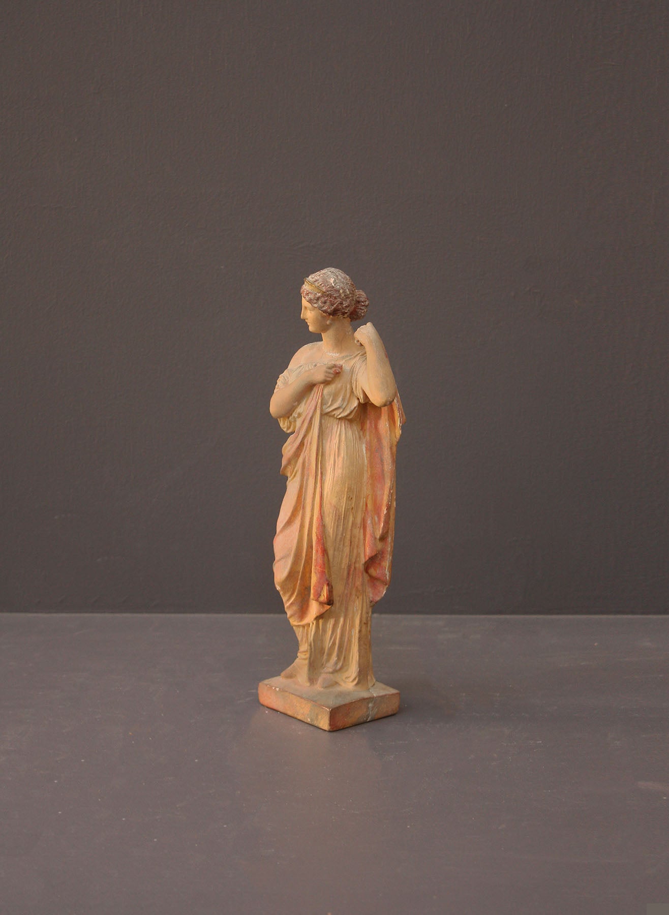 photo of plaster cast statue of female in pink robes with left arm raised and holding robe on gray background