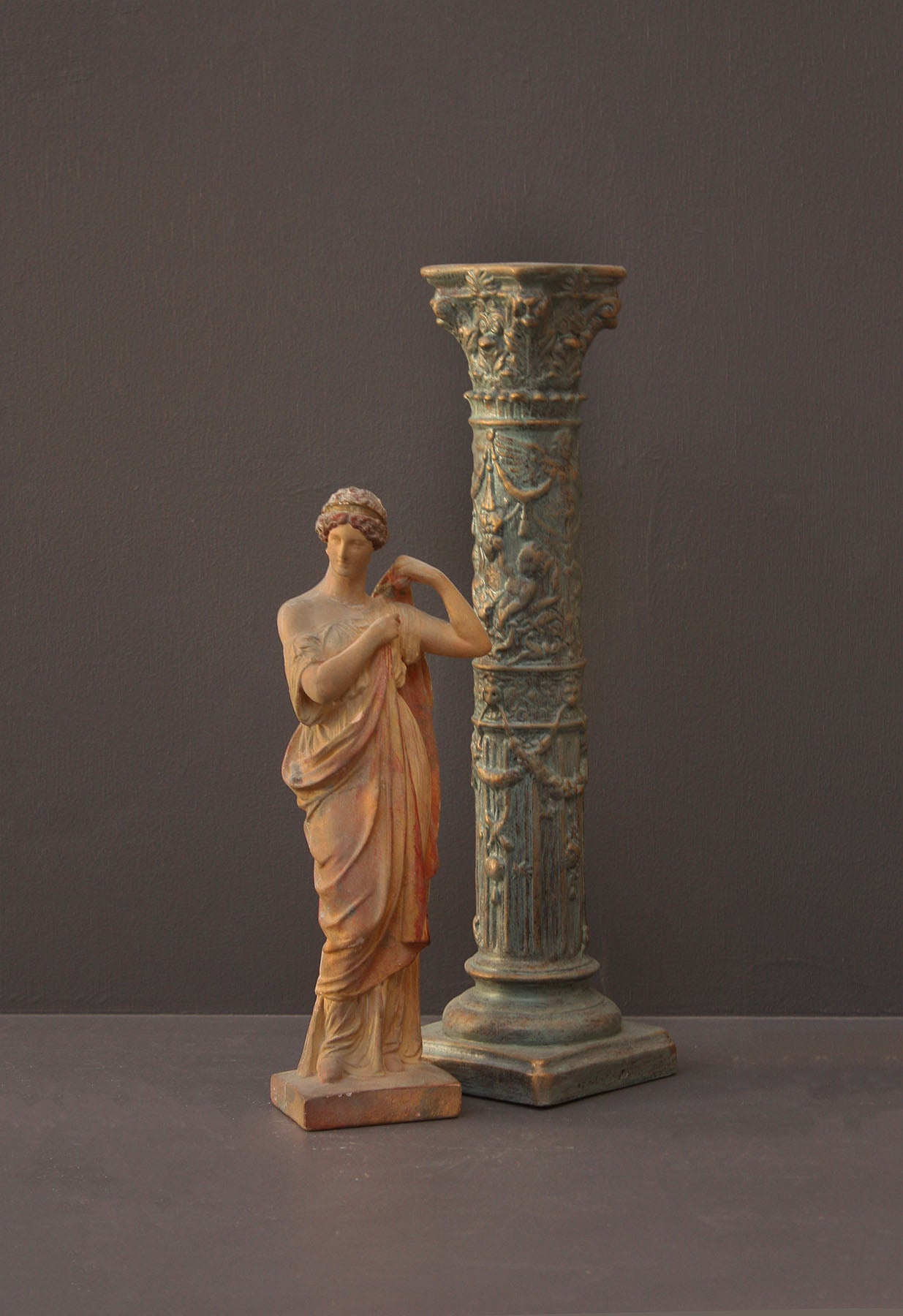 photo of plaster cast statue of female in pink robes with left arm raised and holding robe beside a column-styled candlestick on gray background