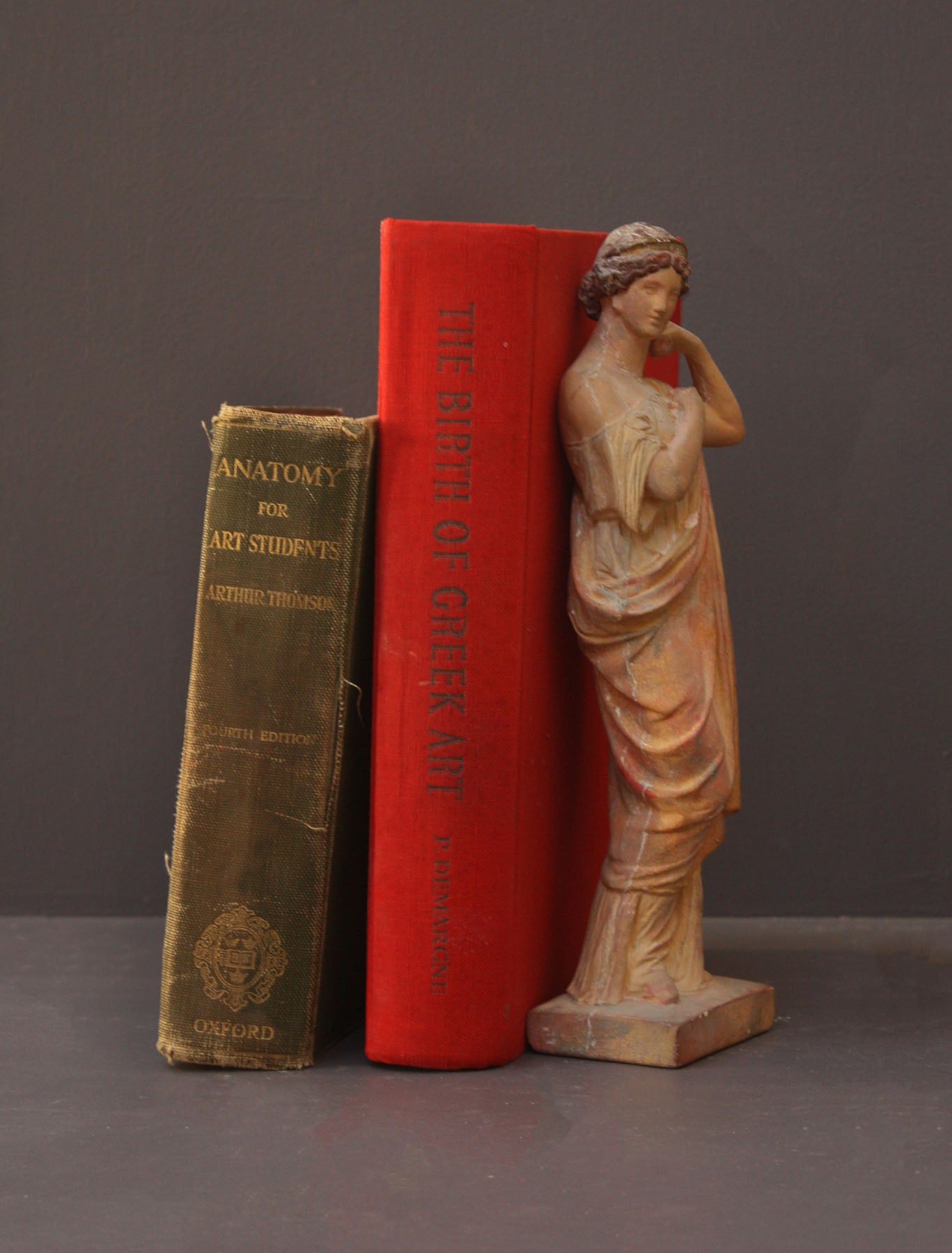 photo of plaster cast statue of female in pink robes with left arm raised and holding robe beside two books on gray background