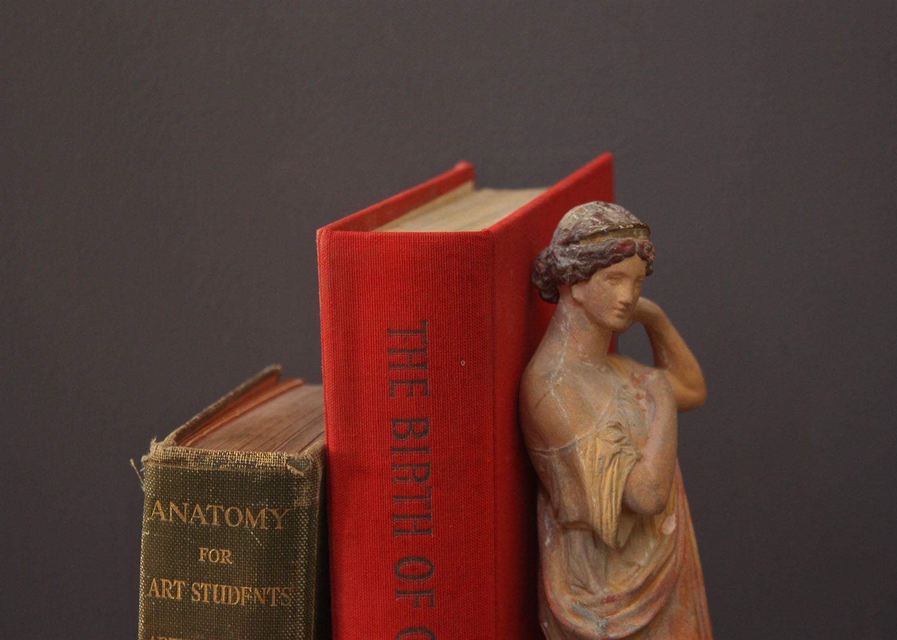 photo of plaster cast statue of female in pink robes with left arm raised and holding robe beside two books on gray background