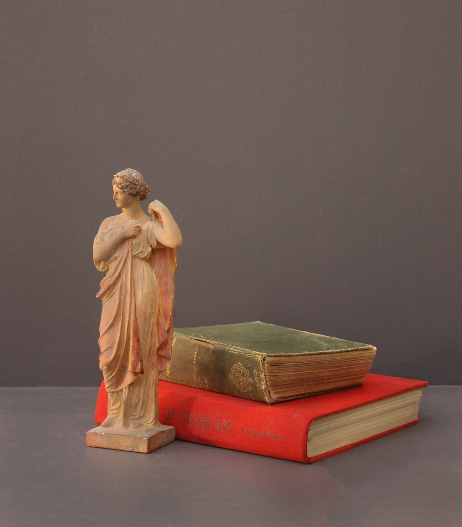 photo of plaster cast statue of female in pink robes with left arm raised and holding robe beside two stacked books on gray background