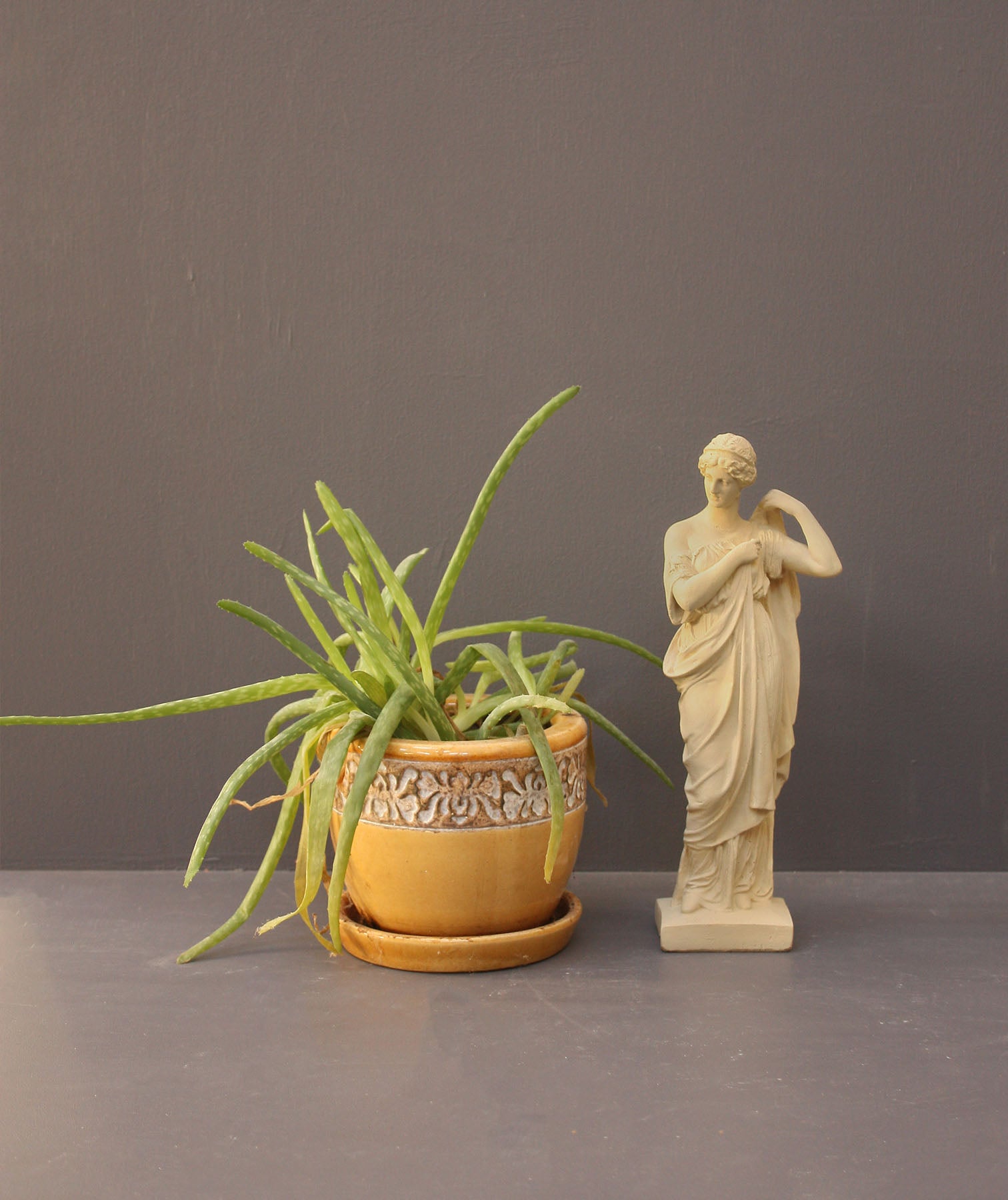 photo of off-white plaster cast statue of female in robes with left arm raised and holding robe beside a potted plant on gray background