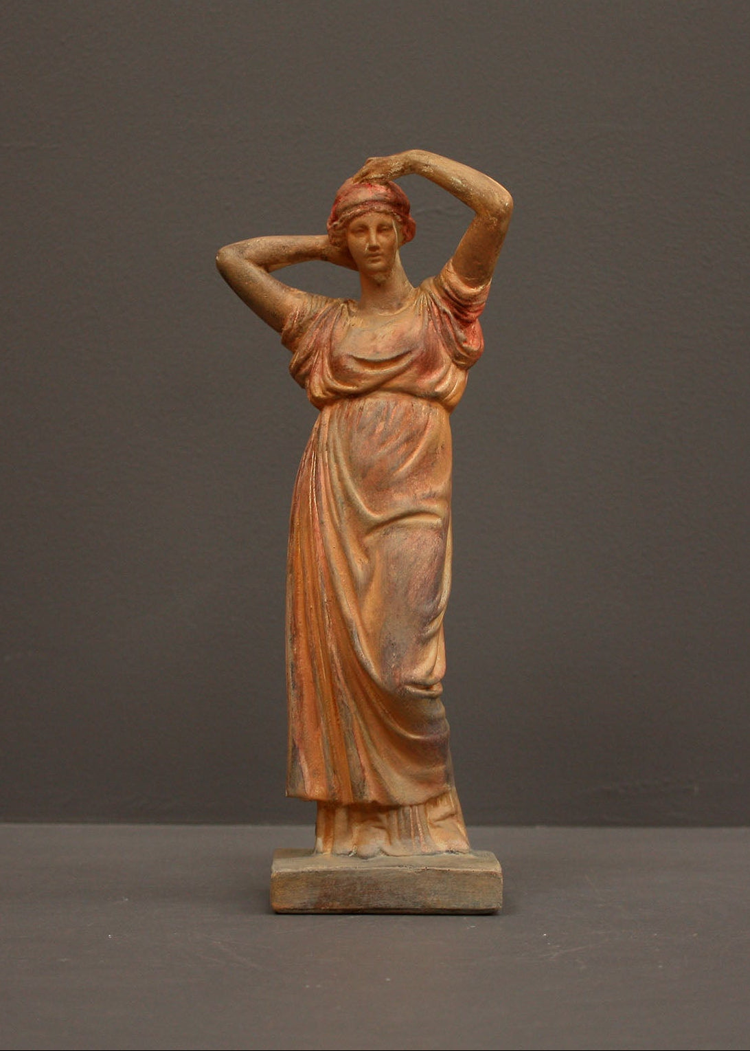photo of plaster cast statue of a female in pink-orange tinted robes with her arms raised to her head against gray background