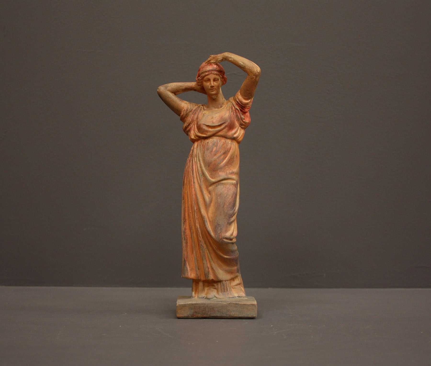 photo of plaster cast statue of a female in pink-orange tinted robes with her arms raised to her head against gray background