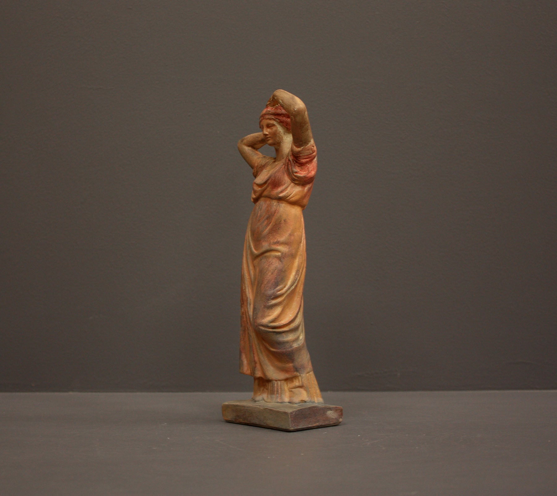 photo of plaster cast statue of a female in pink-orange tinted robes with her arms raised to her head against gray background
