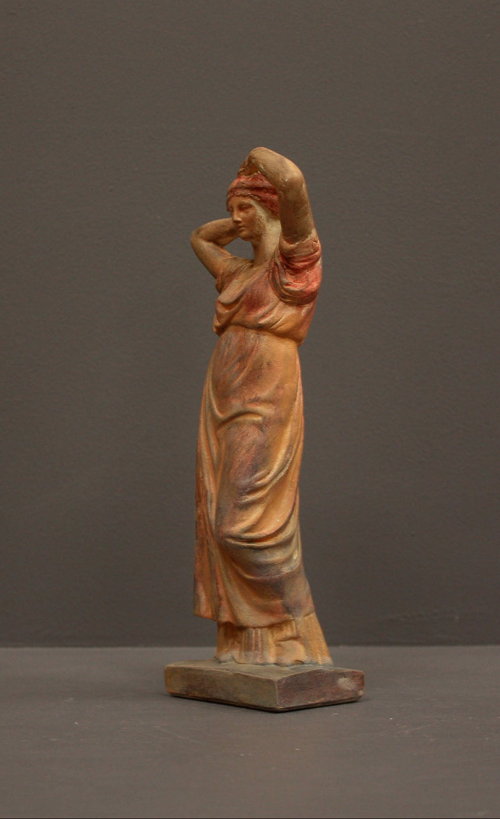 photo of plaster cast statue of a female in pink-orange tinted robes with her arms raised to her head against gray background