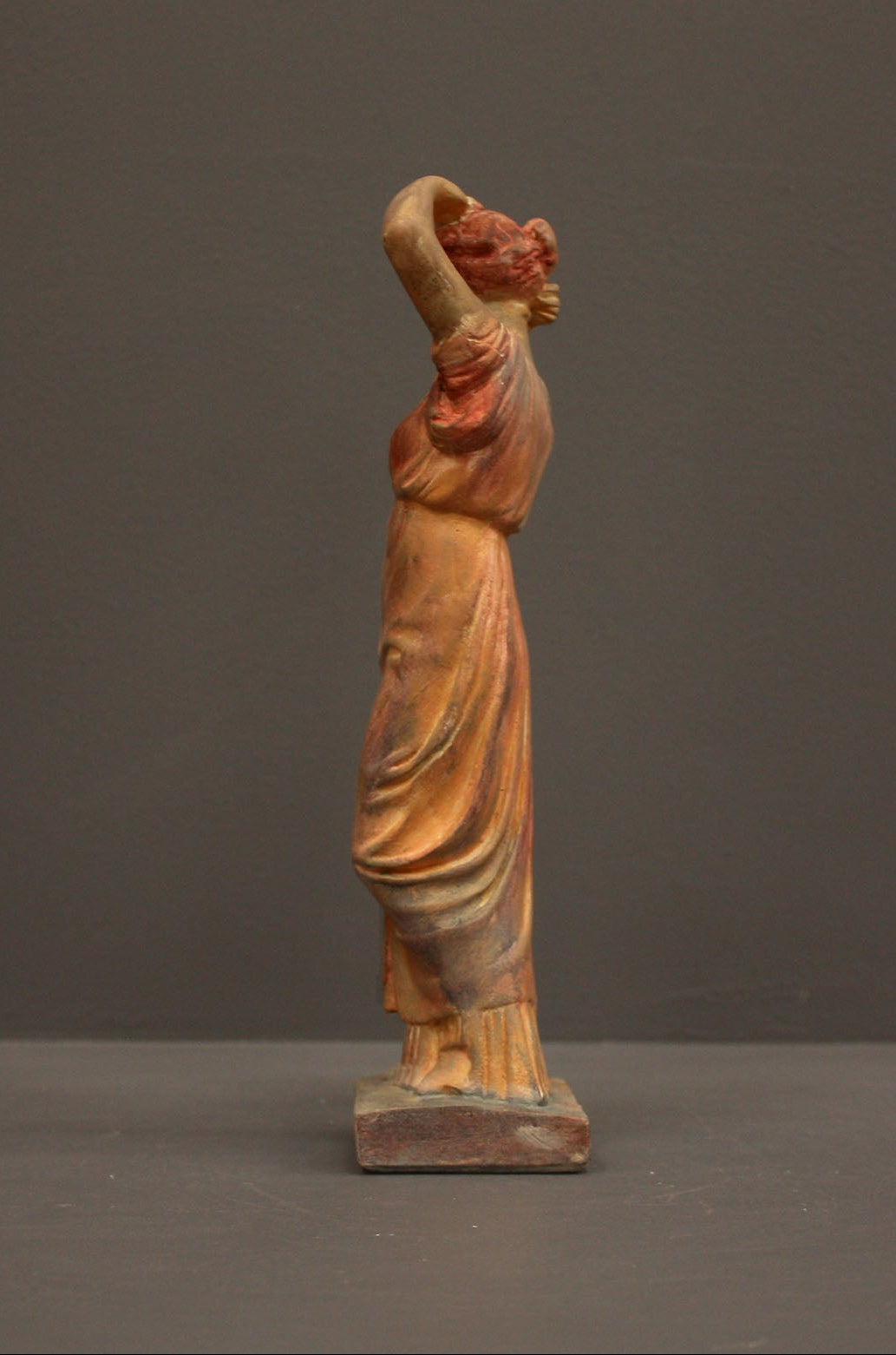 photo of plaster cast statue of a female in pink-orange tinted robes with her arms raised to her head against gray background