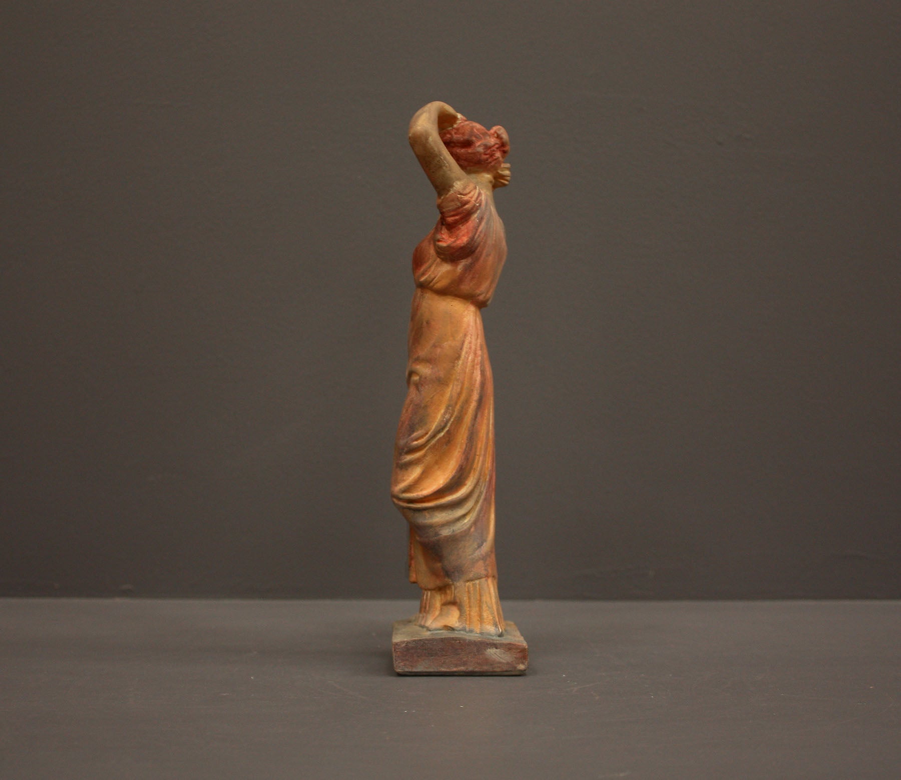 photo of plaster cast statue of a female in pink-orange tinted robes with her arms raised to her head against gray background