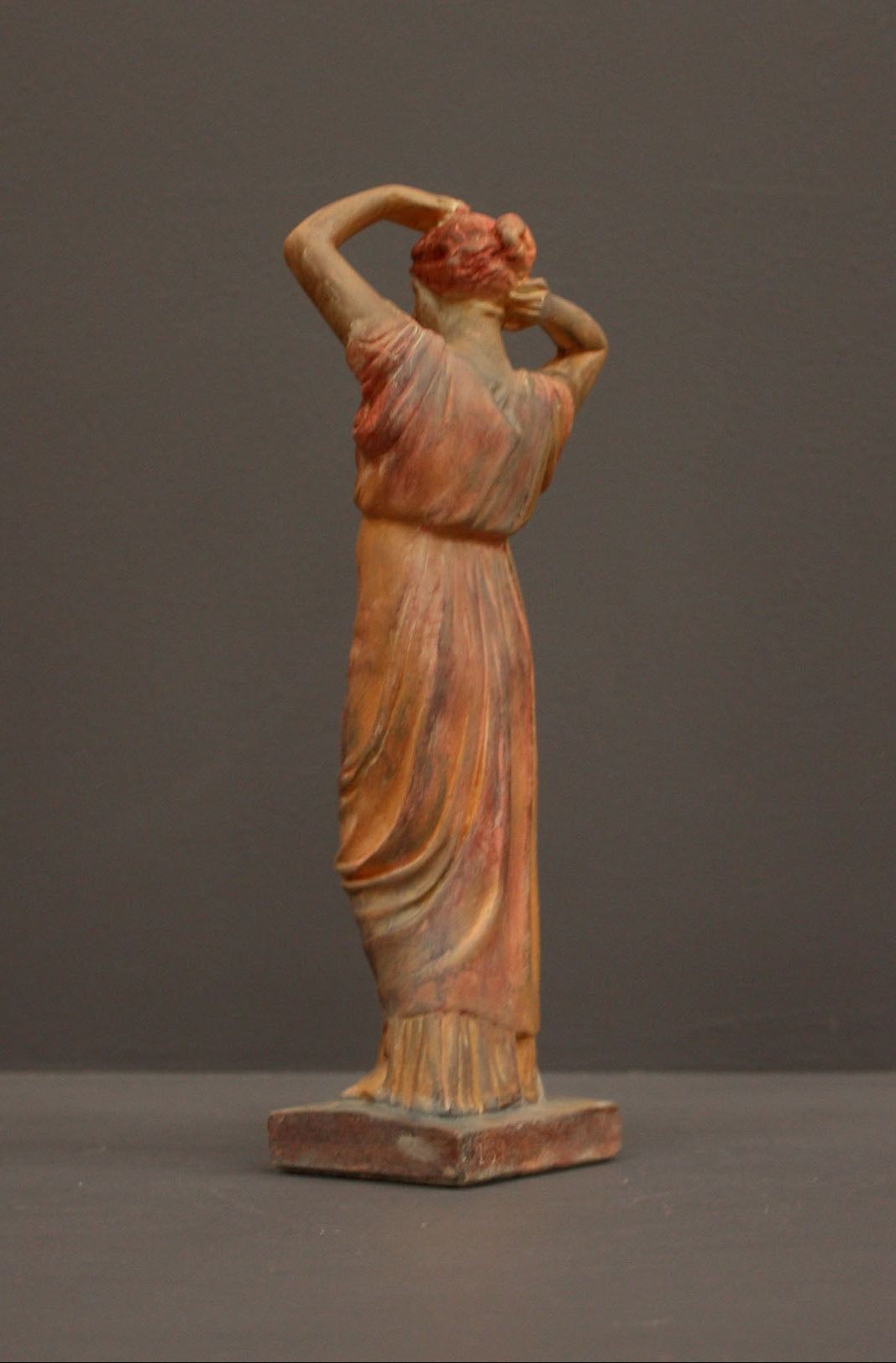 photo of plaster cast statue of a female in pink-orange tinted robes with her arms raised to her head against gray background