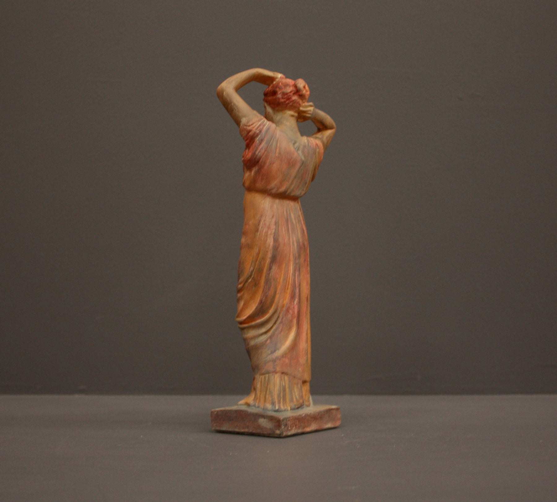 photo of plaster cast statue of a female in pink-orange tinted robes with her arms raised to her head against gray background