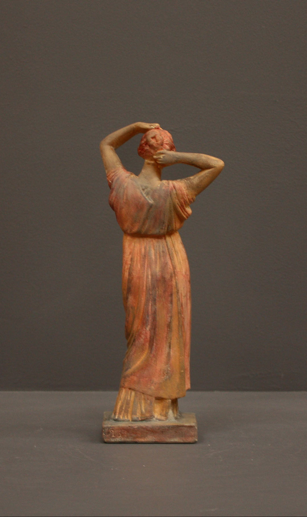 photo of plaster cast statue of a female in pink-orange tinted robes with her arms raised to her head against gray background