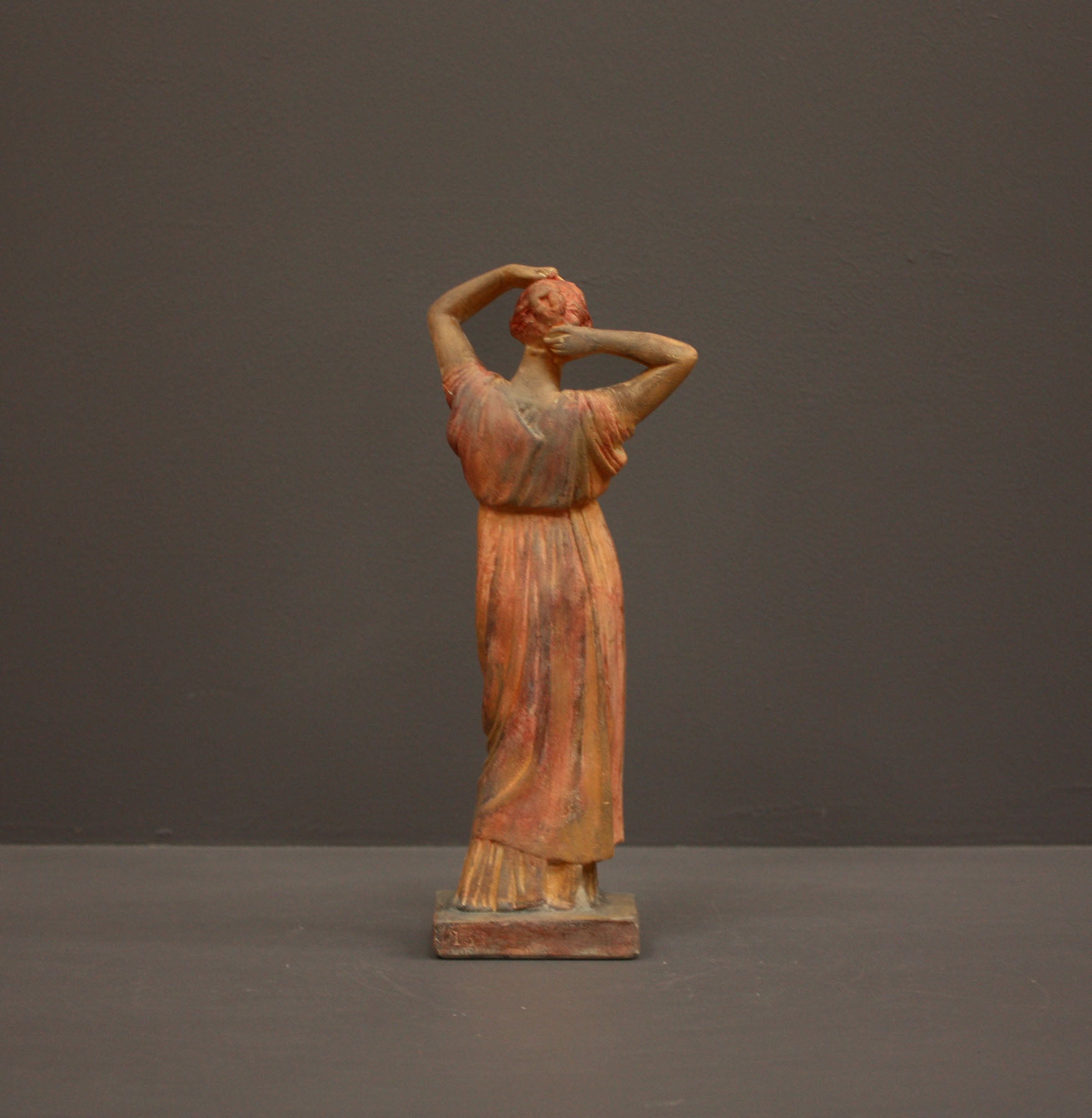 photo of plaster cast statue of a female in pink-orange tinted robes with her arms raised to her head against gray background