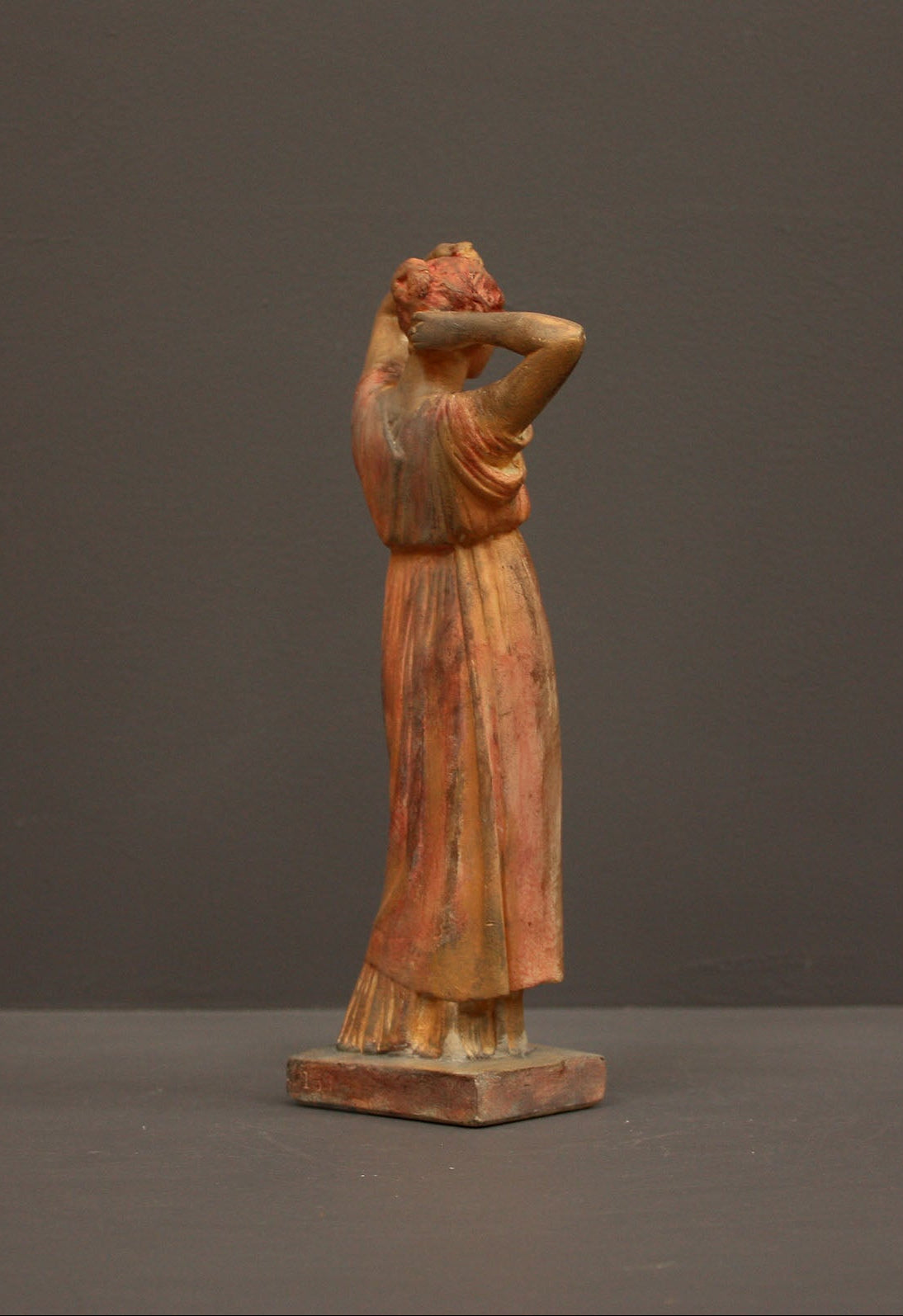 photo of plaster cast statue of a female in pink-orange tinted robes with her arms raised to her head against gray background