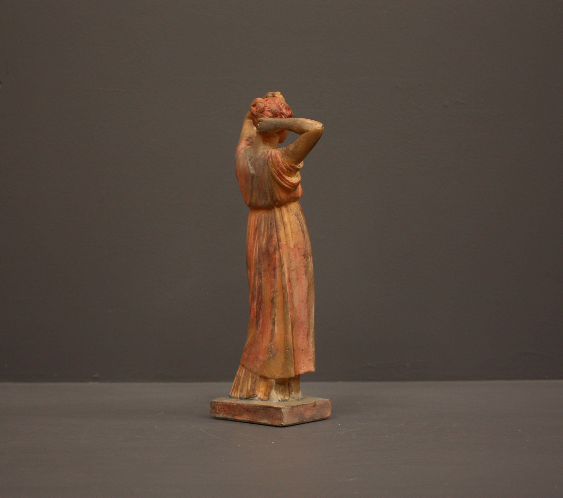 photo of plaster cast statue of a female in pink-orange tinted robes with her arms raised to her head against gray background