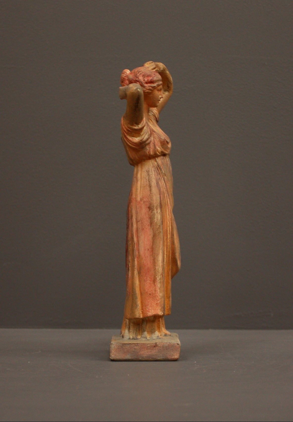 photo of plaster cast statue of a female in pink-orange tinted robes with her arms raised to her head against gray background