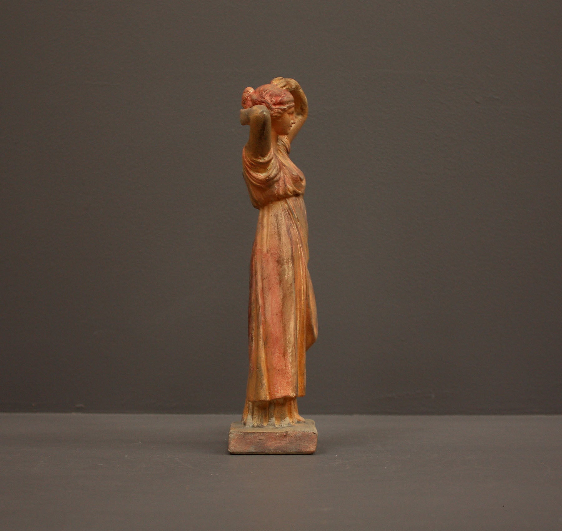 photo of plaster cast statue of a female in pink-orange tinted robes with her arms raised to her head against gray background