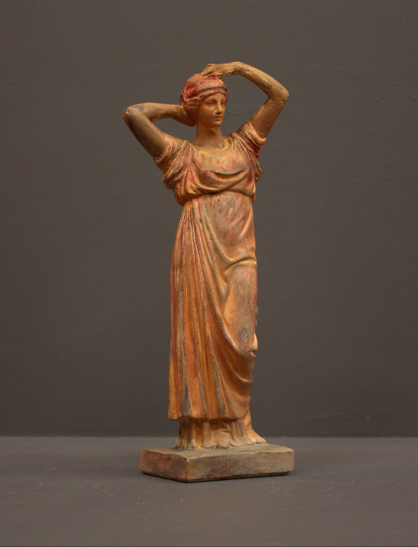 photo of plaster cast statue of a female in pink-orange tinted robes with her arms raised to her head against gray background