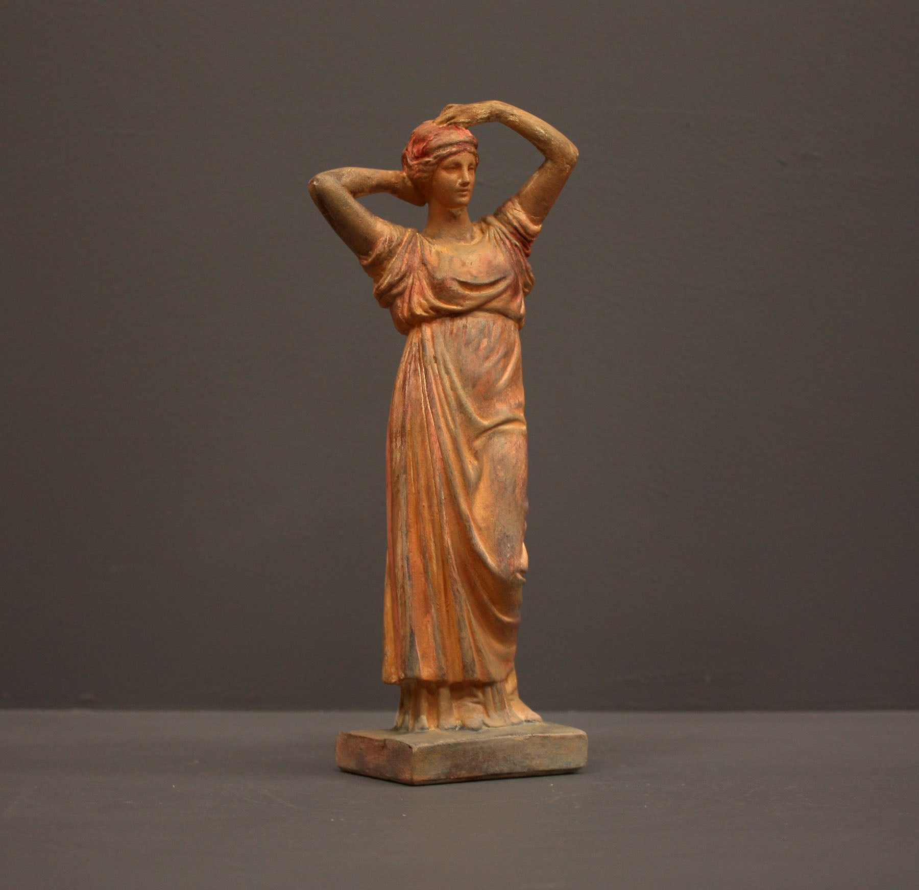 photo of plaster cast statue of a female in pink-orange tinted robes with her arms raised to her head against gray background