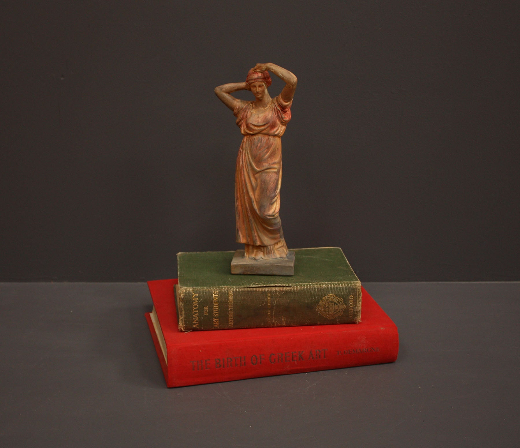 photo of plaster cast statue of a female in pink-orange tinted robes with her arms raised to her head atop green and red books against gray background