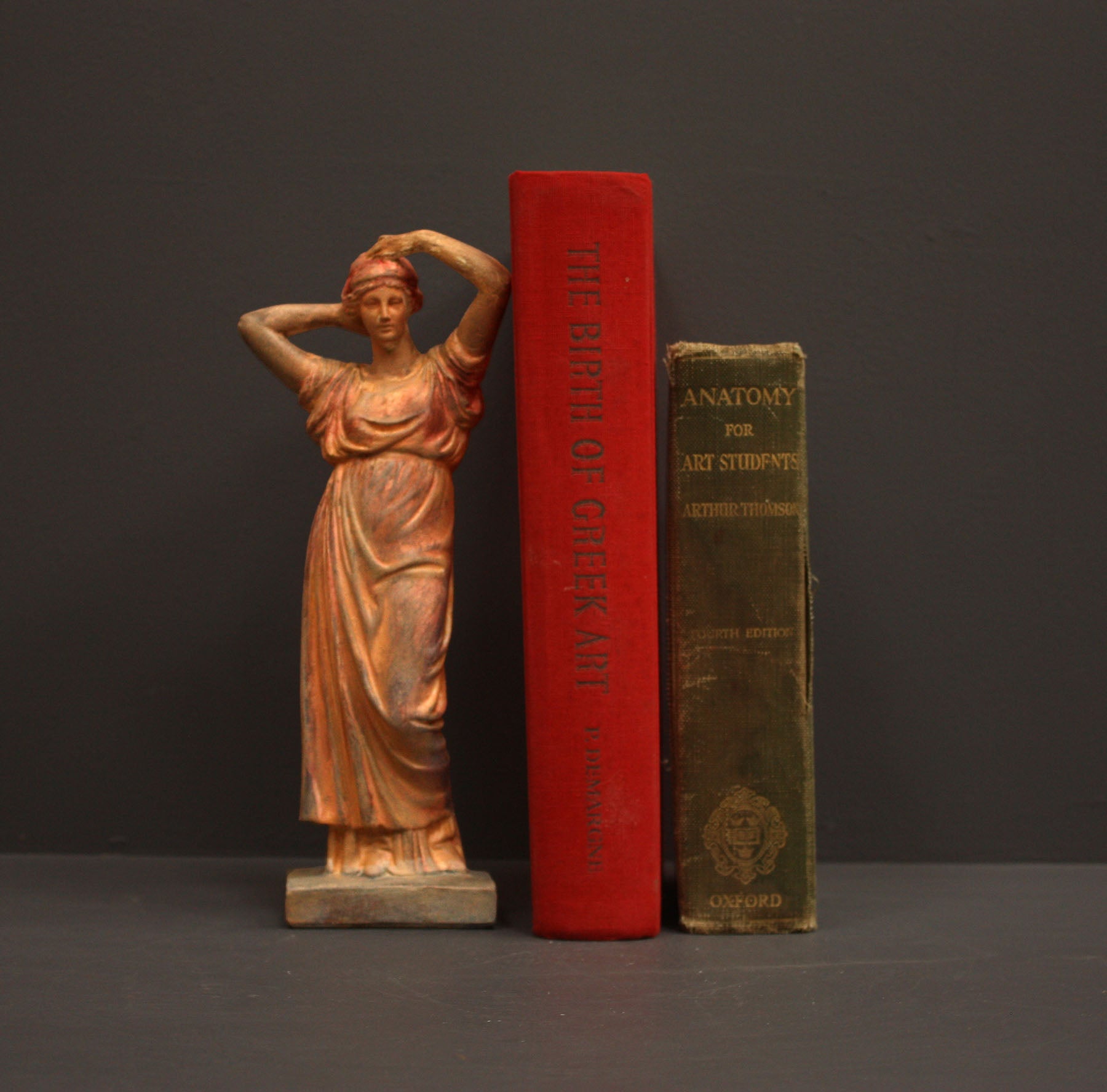 photo of plaster cast statue of a female in pink-orange tinted robes with her arms raised to her head beside red and green books against gray background