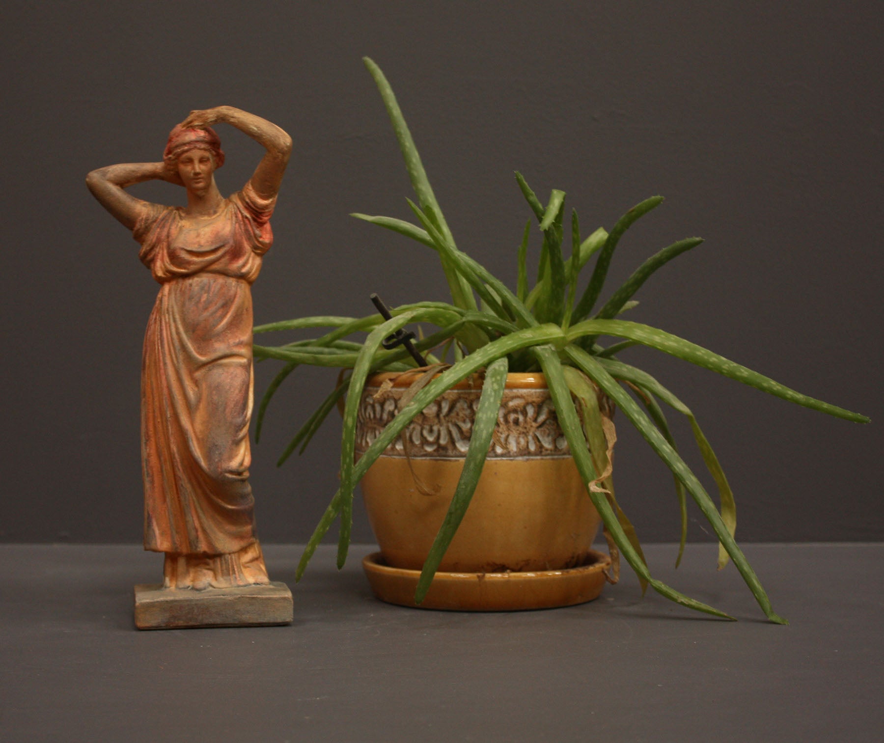 photo of plaster cast statue of a female in pink-orange tinted robes with her arms raised to her head beside potted plant against gray background