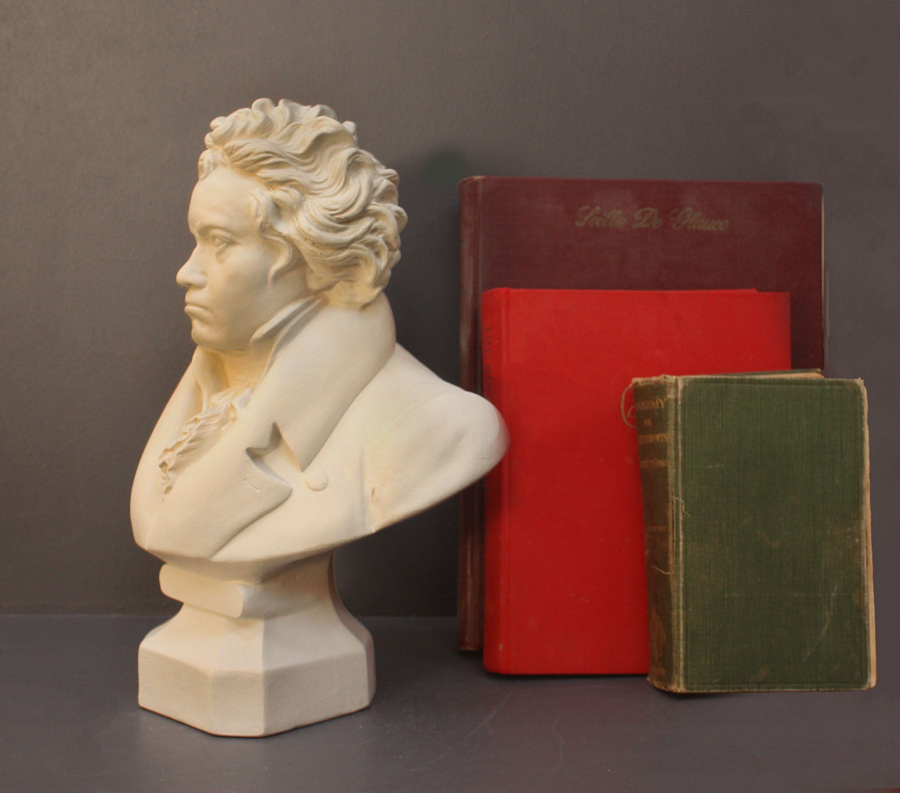photo of plaster cast sculpture of male bust of Ludwig van Beethoven in dress coat and ruffled necktie set beside green and red books on gray background