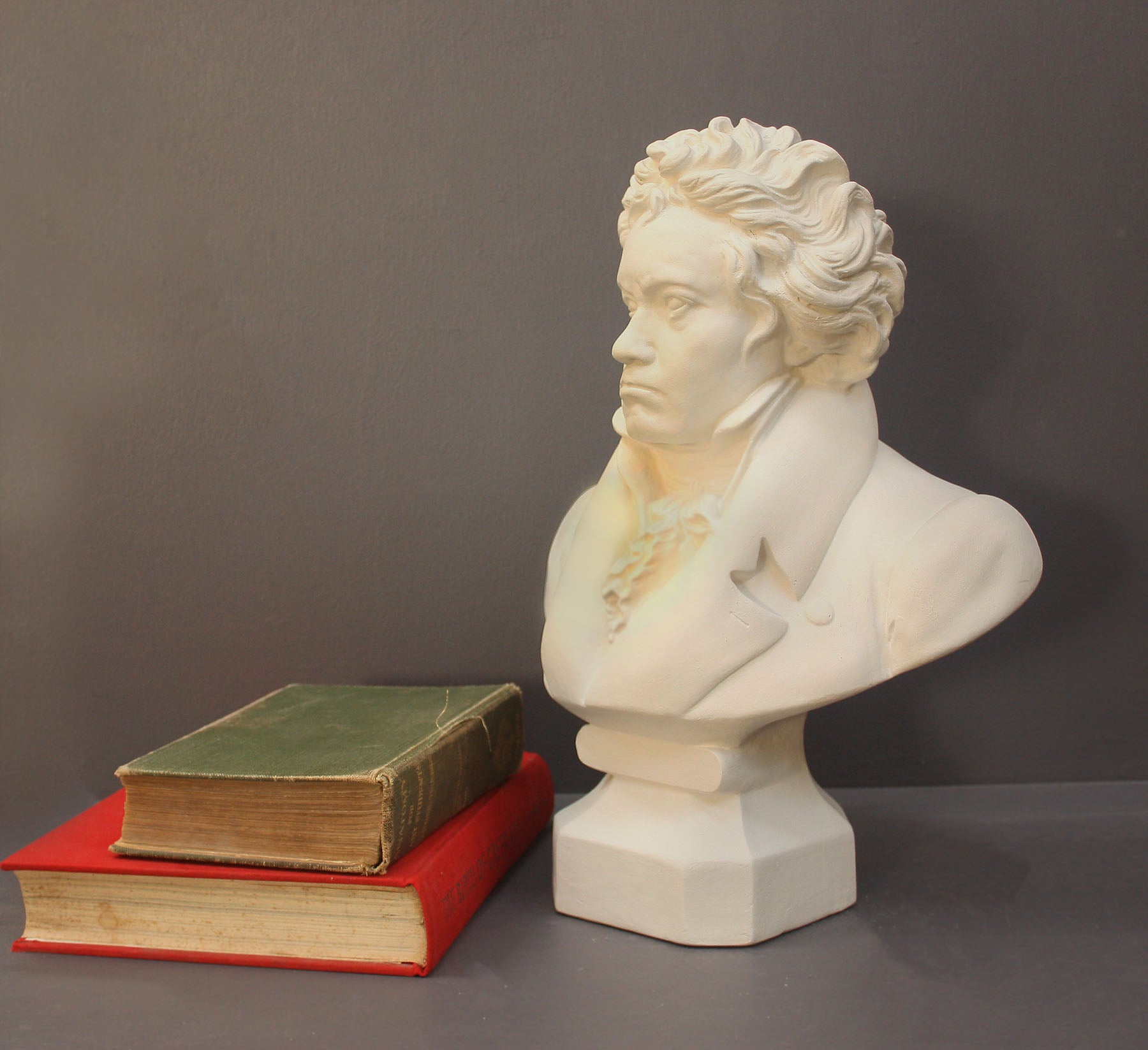 photo of plaster cast sculpture of male bust of Ludwig van Beethoven in dress coat and ruffled necktie set beside green and red books on gray background