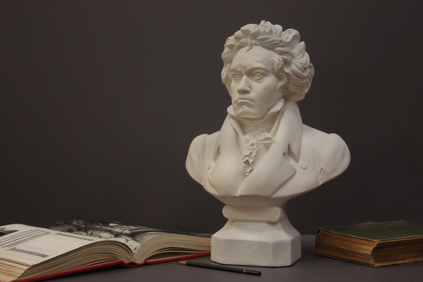 photo of plaster cast sculpture of male bust of Ludwig van Beethoven in dress coat and ruffled necktie set beside an open book to the left, a closed green book to the right, and a black pencil in front of it on gray background