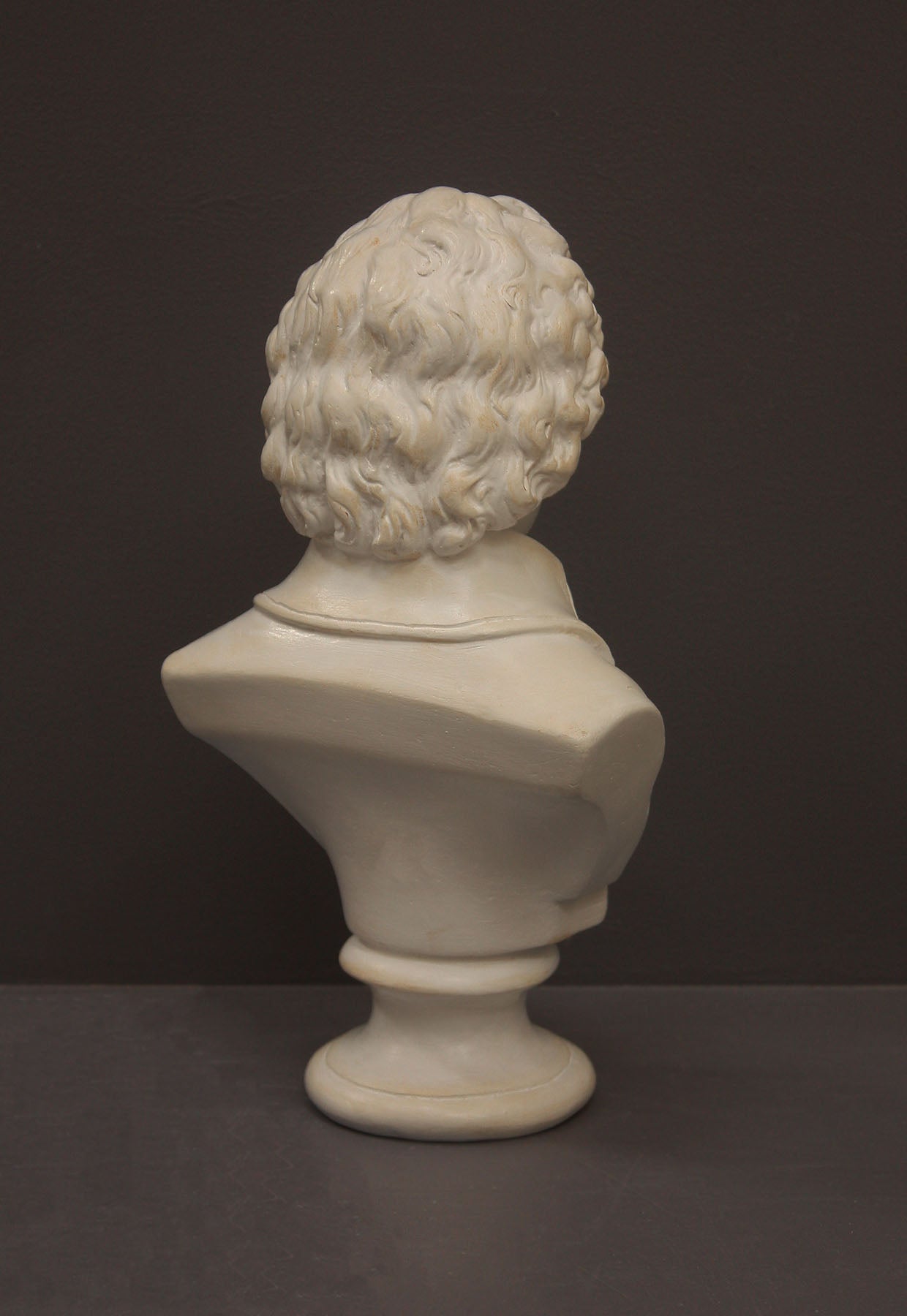 back view image of a plaster sculpture bust of man, namely Mozart, with ruffled necktie and coat on a gray background