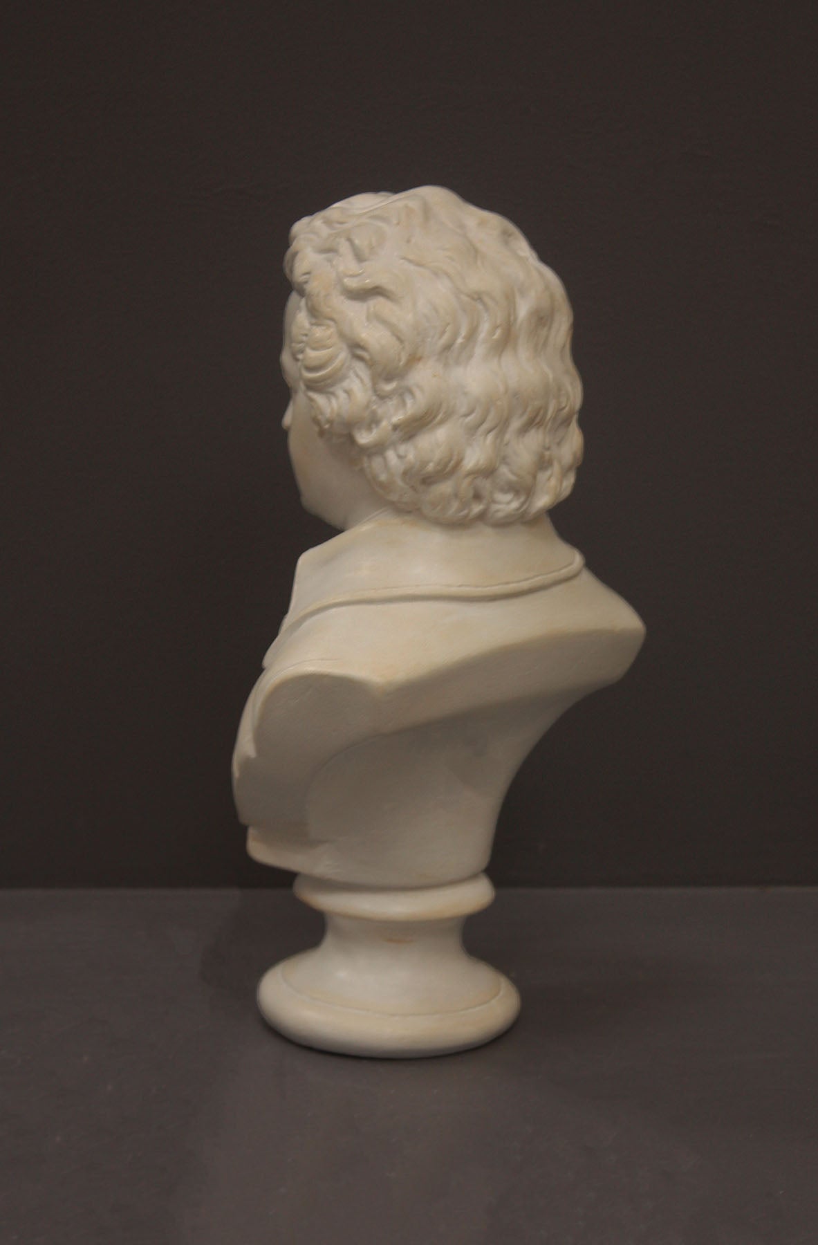 back view image of a plaster sculpture bust of man, namely Mozart, with ruffled necktie and coat on a gray background
