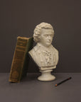 image of a plaster sculpture bust of man, namely Mozart, with ruffled necktie and coat beside a black drawing pencil and with a green book leaning against the bust on a gray background