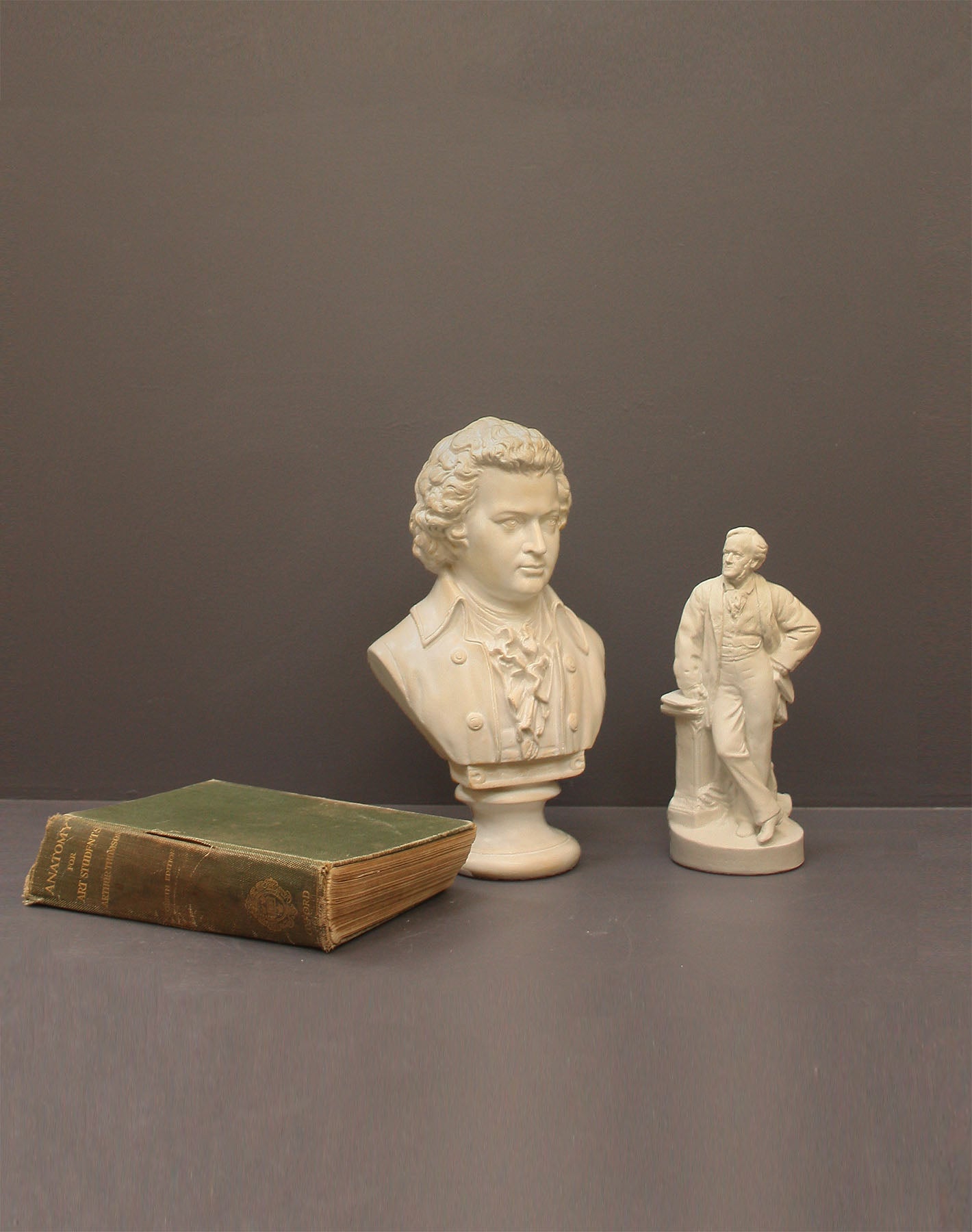 image of a plaster sculpture bust of man, namely Mozart, with ruffled necktie and coat and smaller statue of a man, namely Wagner, with hand on hip leaning against a podium and with a green book beside the sculptures on a gray background