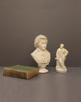 image of a plaster sculpture bust of man, namely Mozart, with ruffled necktie and coat and smaller statue of a man, namely Wagner, with hand on hip leaning against a podium and with a green book beside the sculptures on a gray background