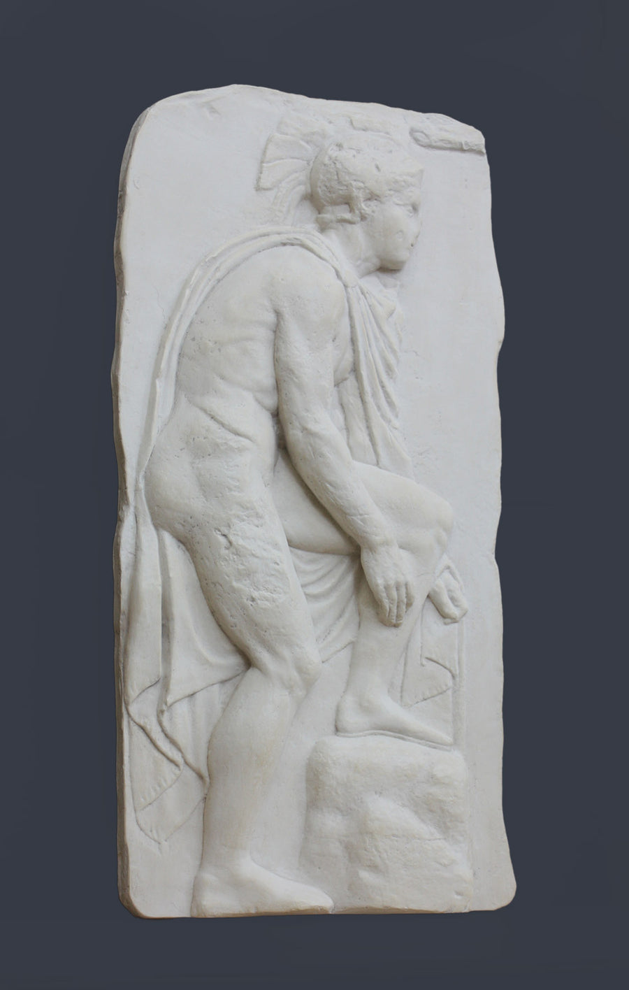 photo of off-white plaster cast relief sculpture of male nude with cloak bending to tie his sandal against gray background
