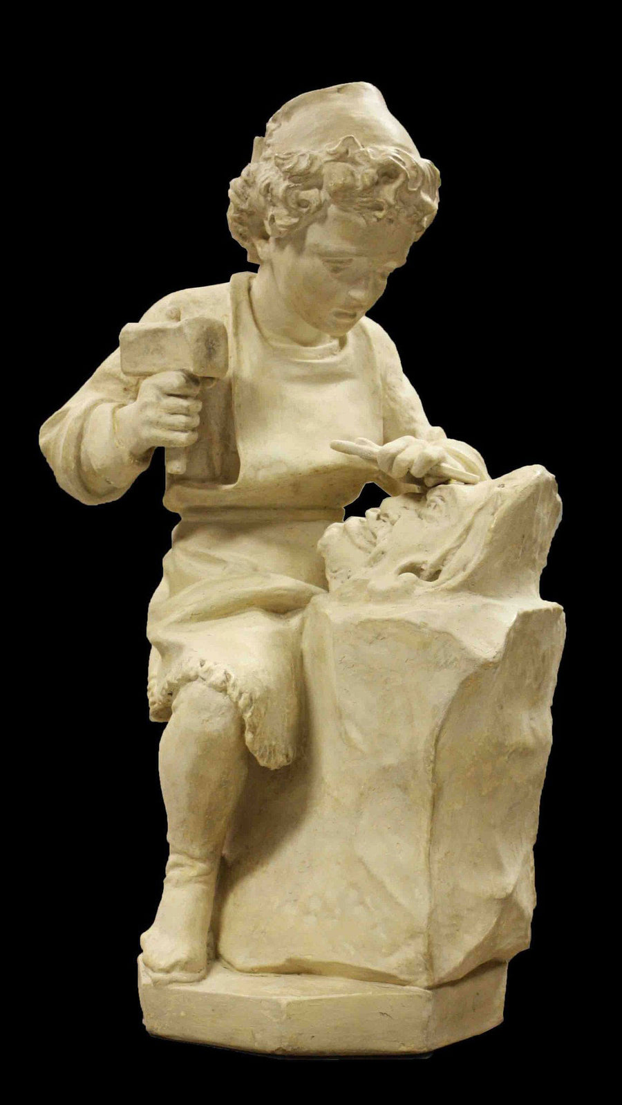 photo with black background of plaster cast of child, namely Michelangelo, on stool with chisel and hammer in hand and sculpting a faun mask from a block of stone